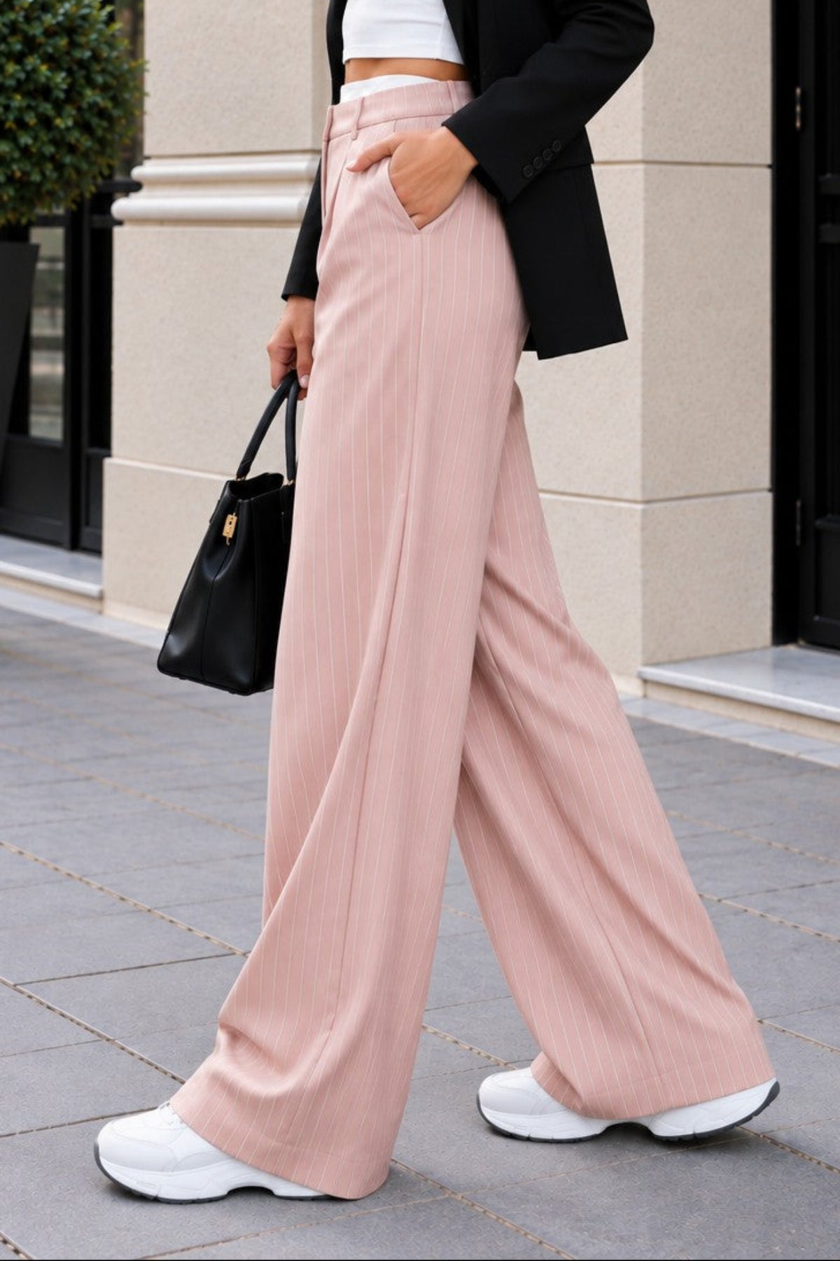 A woman walking outdoors in a city setting wearing a black blazer, white crop top, and dusty pink wide-leg pants, carrying a black handbag.