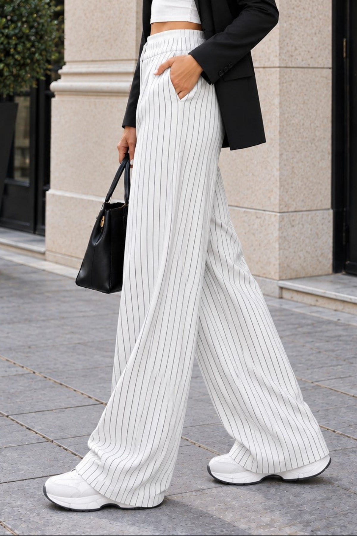 A woman walking outdoors in a city setting wearing a white crop top, white wide-leg pinstriped trousers, a black blazer, white sneakers, and carrying a black handbag.