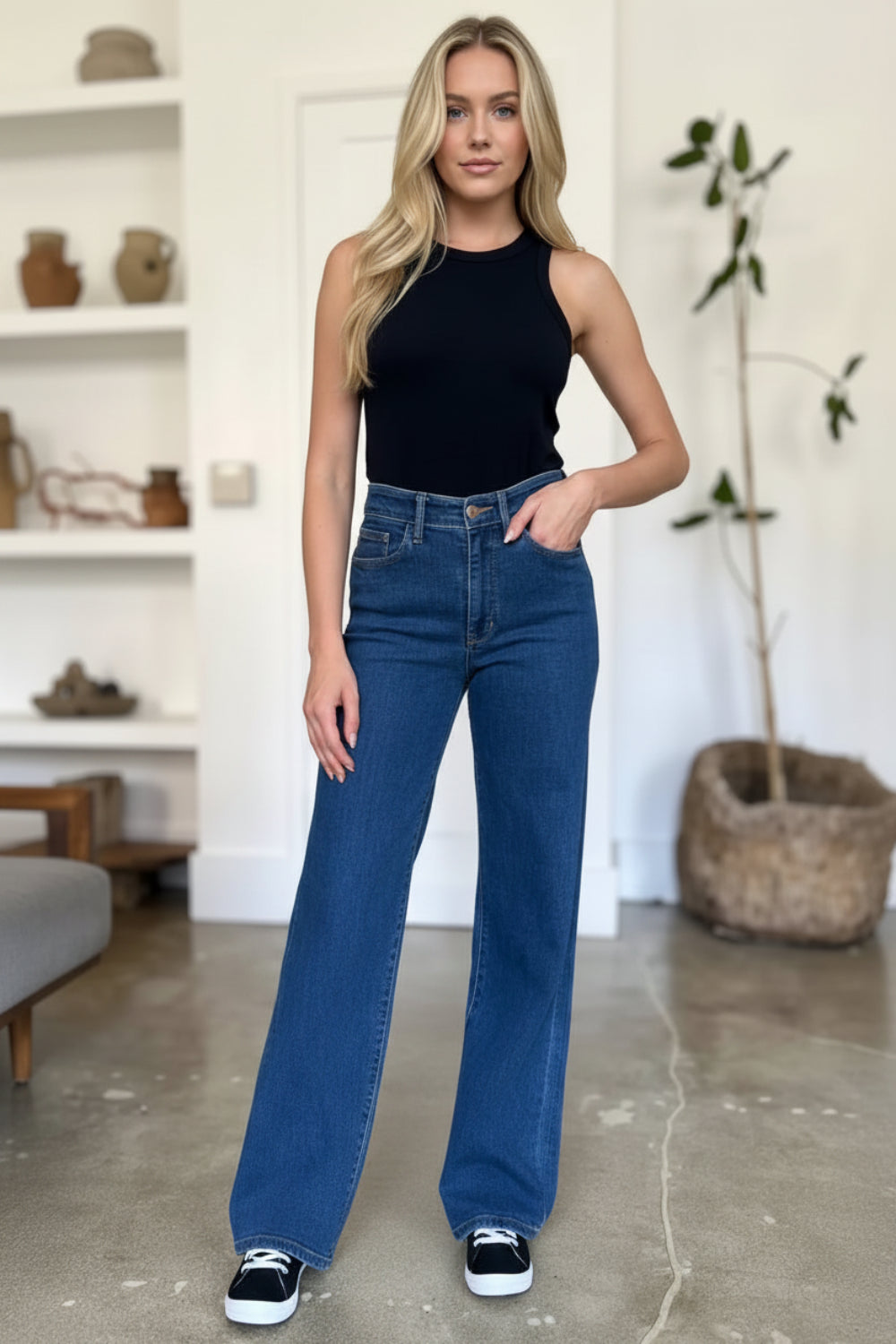 A woman modeling a pair of high-waisted, wide-leg blue jeans with a black sleeveless top and black sneakers, standing in a bright, modern room with shelves and a potted plant.