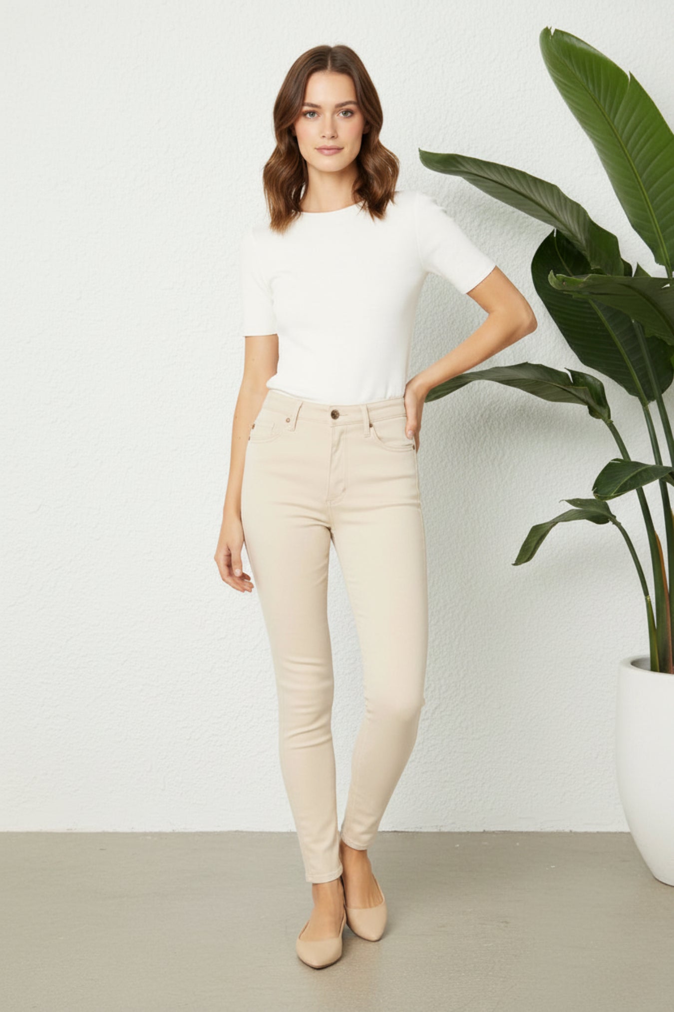 A woman stands in a bright indoor setting wearing a fitted white short-sleeve top, beige ankle-length skinny jeans, and beige flats. A large green potted plant is visible on the right against a white wall.