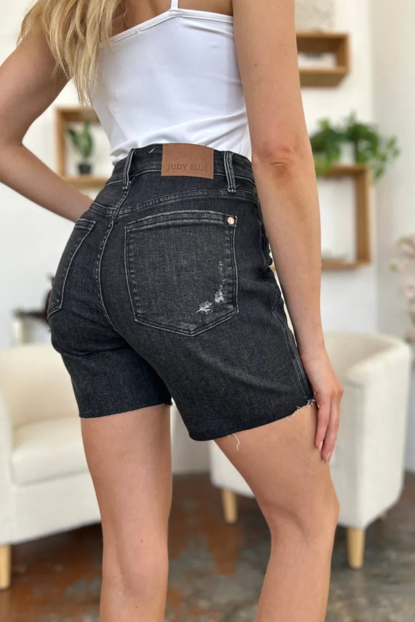 A person wearing dark gray washed denim shorts with a distressed back pocket and a brown leather brand patch, shown from the back in an indoor room setting.