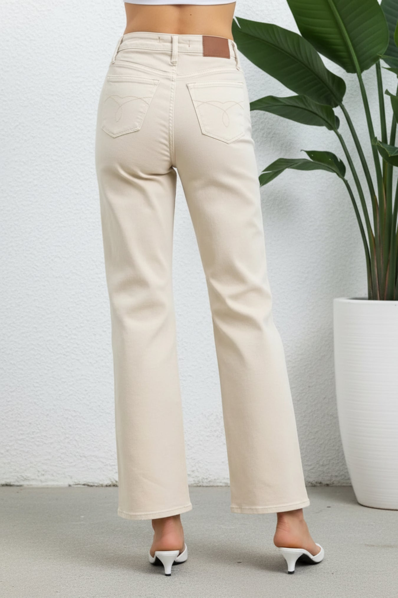A person shown from the back wearing light beige straight-leg jeans with a small brown leather patch at the back waistband, standing indoors against a white wall next to a large potted plant.