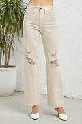 A person wearing light beige wide-leg distressed jeans with ripped details on the thighs, paired with white pointed-toe heels, standing against a white textured wall next to a potted plant.
