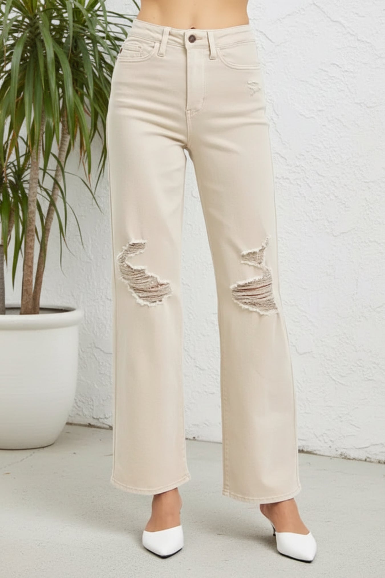 A person wearing light beige wide-leg distressed jeans with ripped details on the thighs, paired with white pointed-toe heels, standing against a white textured wall next to a potted plant.