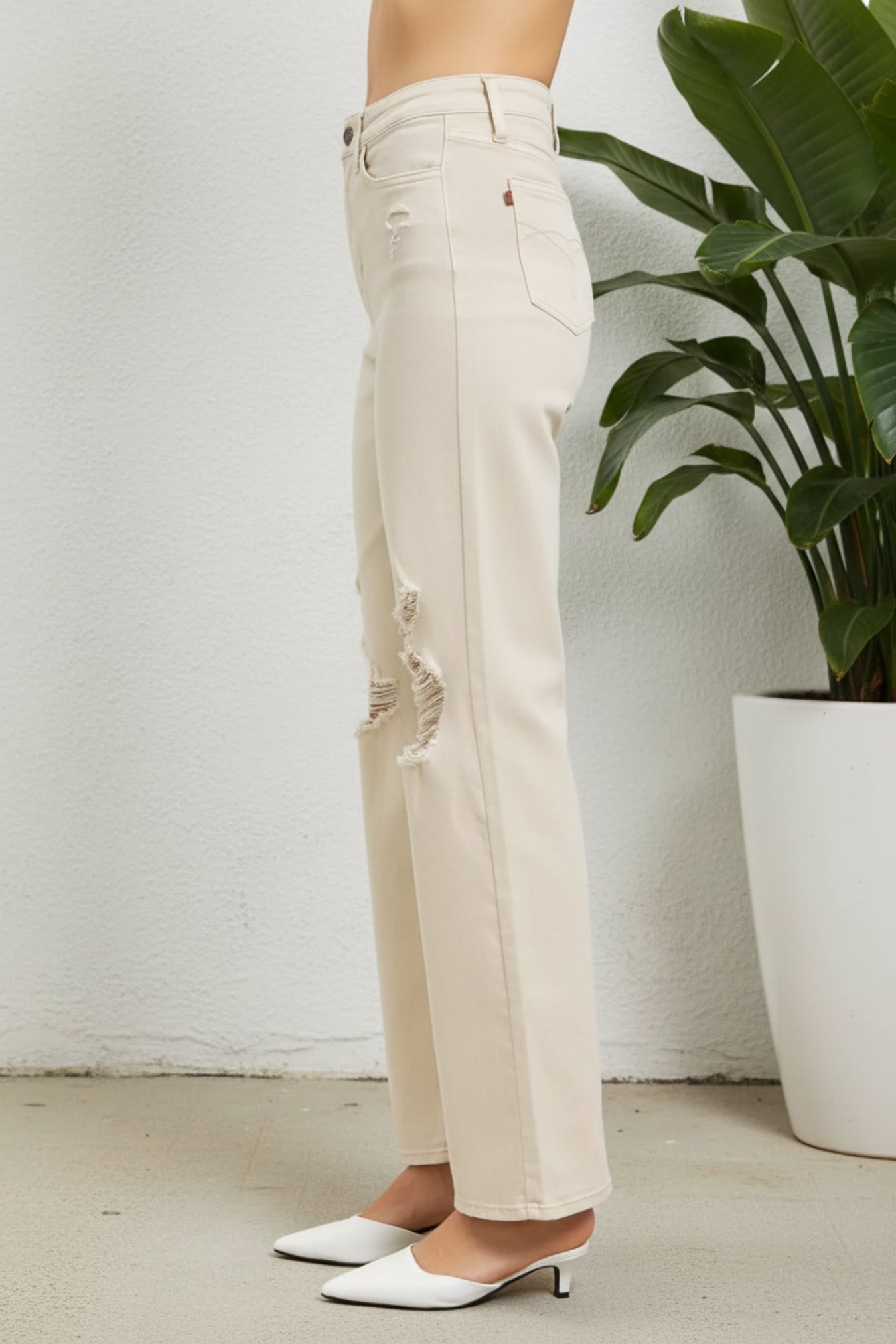 A person wearing off-white distressed jeans with ripped details on the thighs and knees, paired with white pointed-toe mule heels, standing in a room with a white wall and a potted green plant.