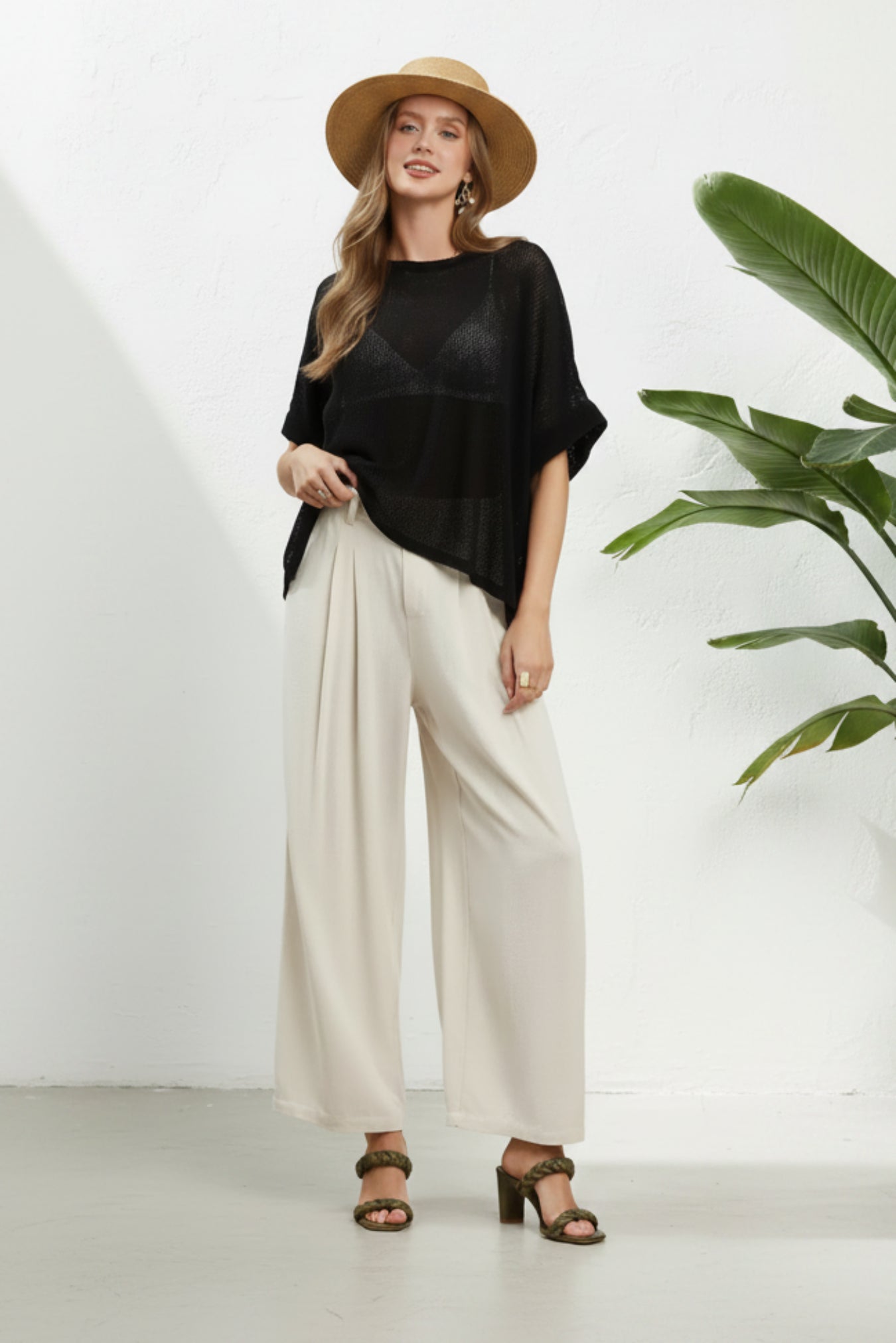 A woman modeling a black short-sleeve top and cream wide-leg pants against a white background, wearing a straw hat and brown sandals next to a white potted plant.