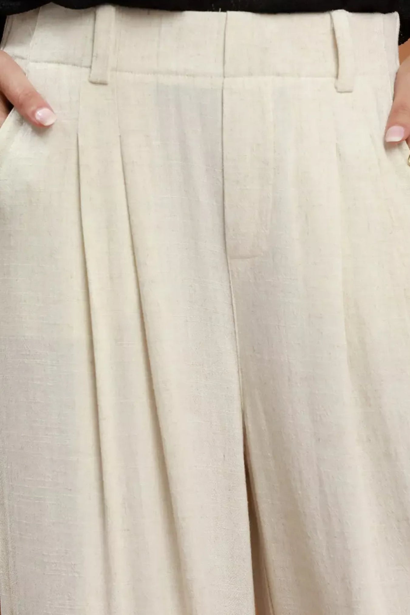 Close-up of a person wearing high-waisted oatmeal wide-leg trousers, showing the fabric texture, pleats, belt loops, and side pockets. The model's hands with manicured nails are visible at the sides, and a black top is partially visible at the top edge.