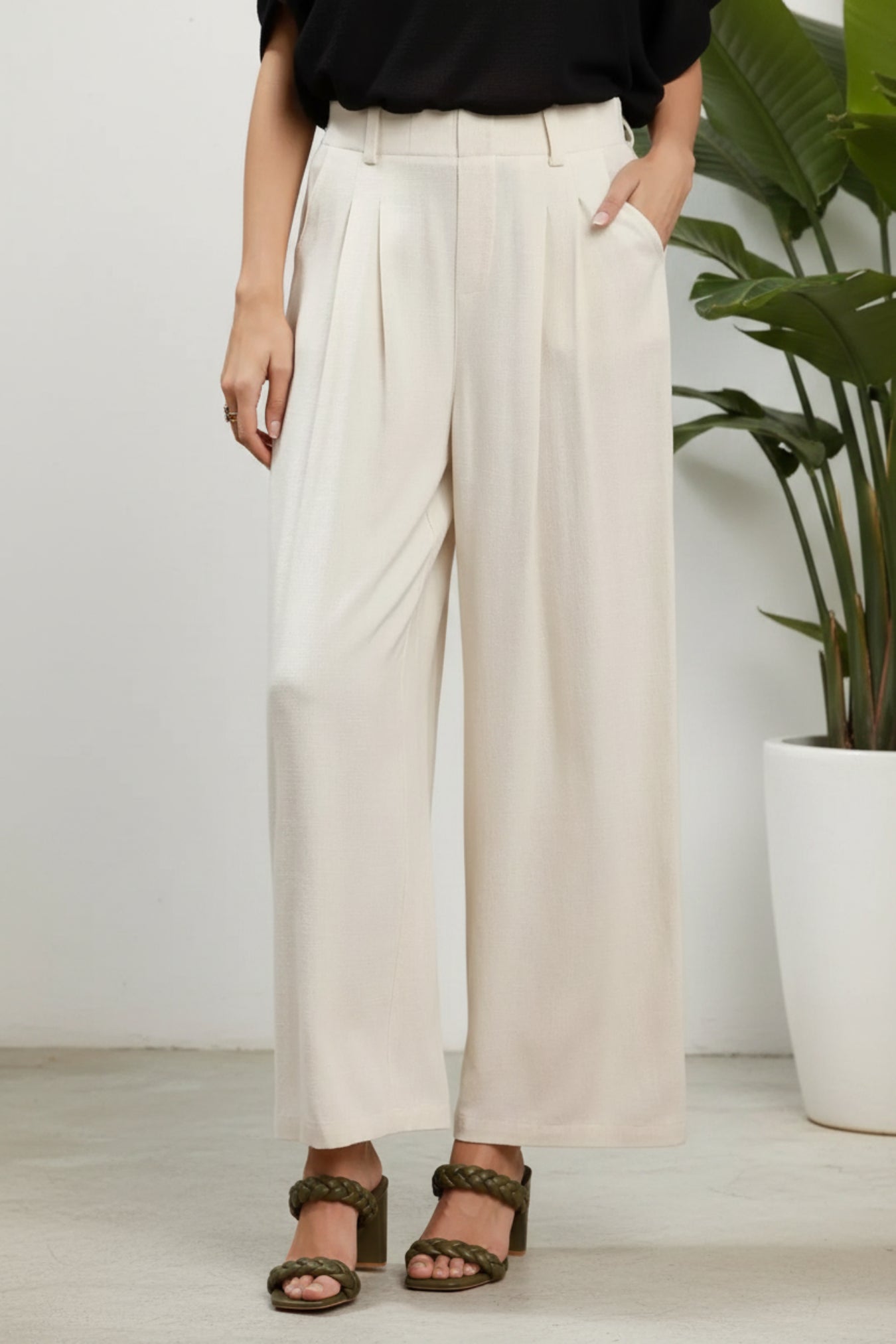 Person wearing oatmeal-colored wide-leg pants on a white background next to a potted plant