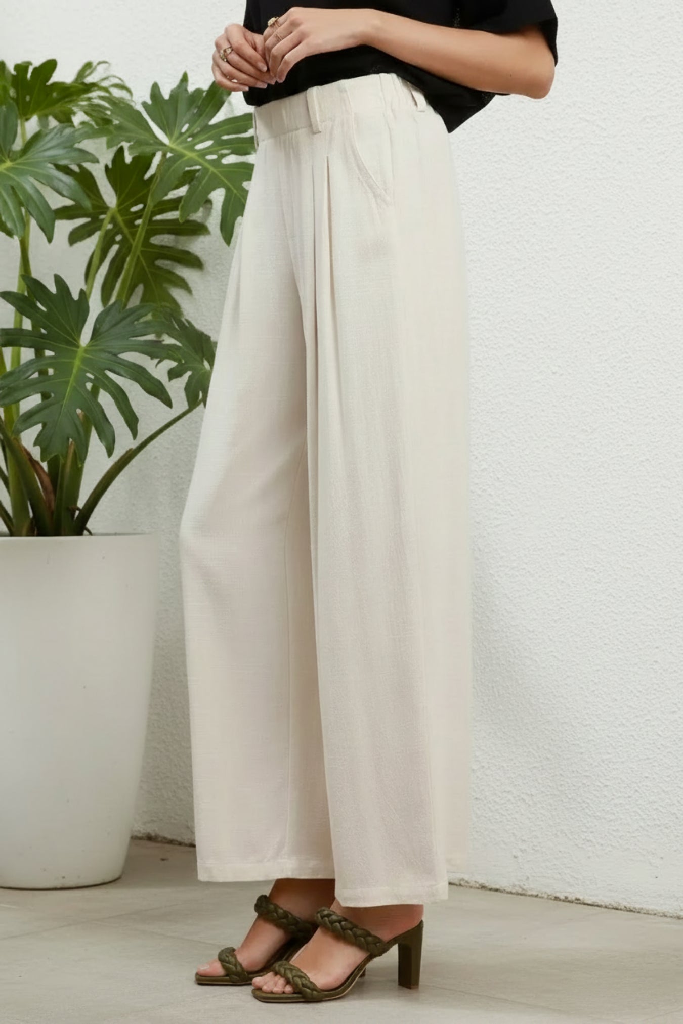 A person wearing high-waisted, wide-leg oatmeal linen pants with a black top and brown heeled sandals, standing against a white background next to a plant.