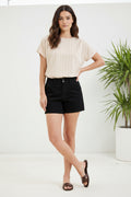 Woman wearing a beige top and black shorts standing in a minimalistic room with a plant.