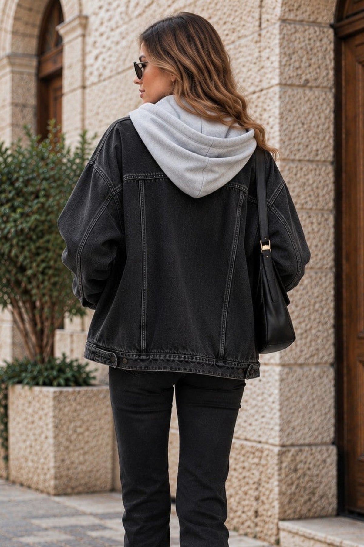 A woman is shown from behind wearing a black denim jacket over a light gray hoodie, black flared jeans, and white sneakers, walking on a cobblestone street next to a stone building.