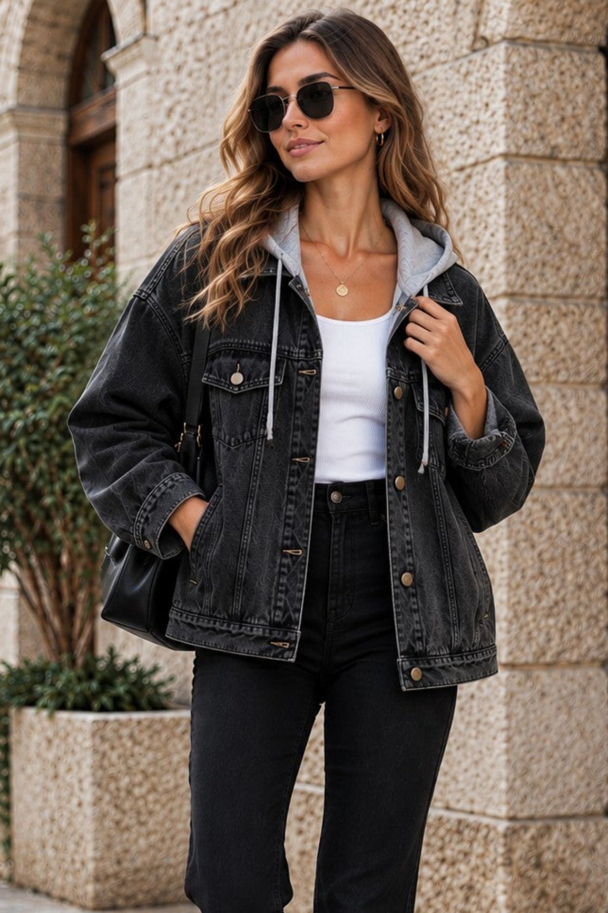 A woman walking on a stone street wearing a black washed denim jacket with a gray hood, a white top, black pants, and white sneakers.
