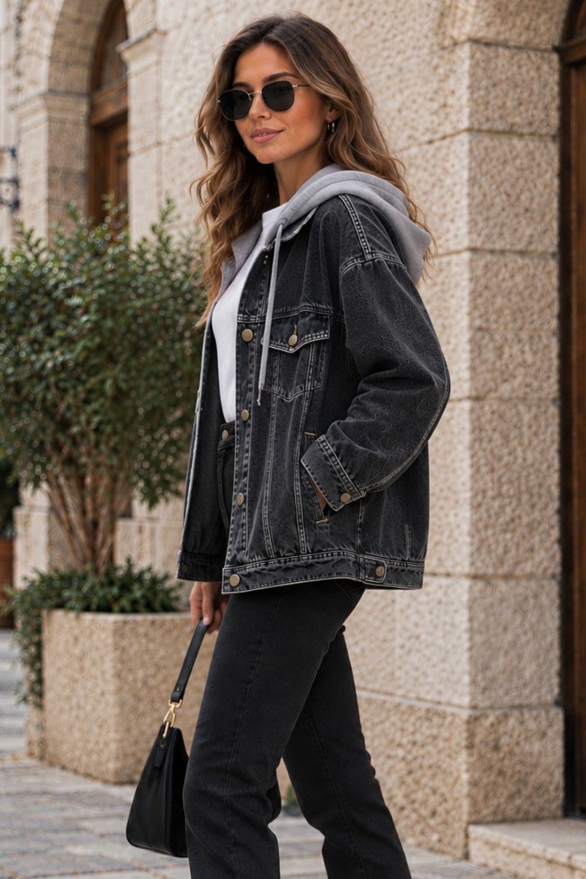 A woman stands on a cobblestone street in front of a stone building, wearing a dark washed denim jacket over a light gray hoodie, black flared jeans, white sneakers, and carrying a small black handbag.