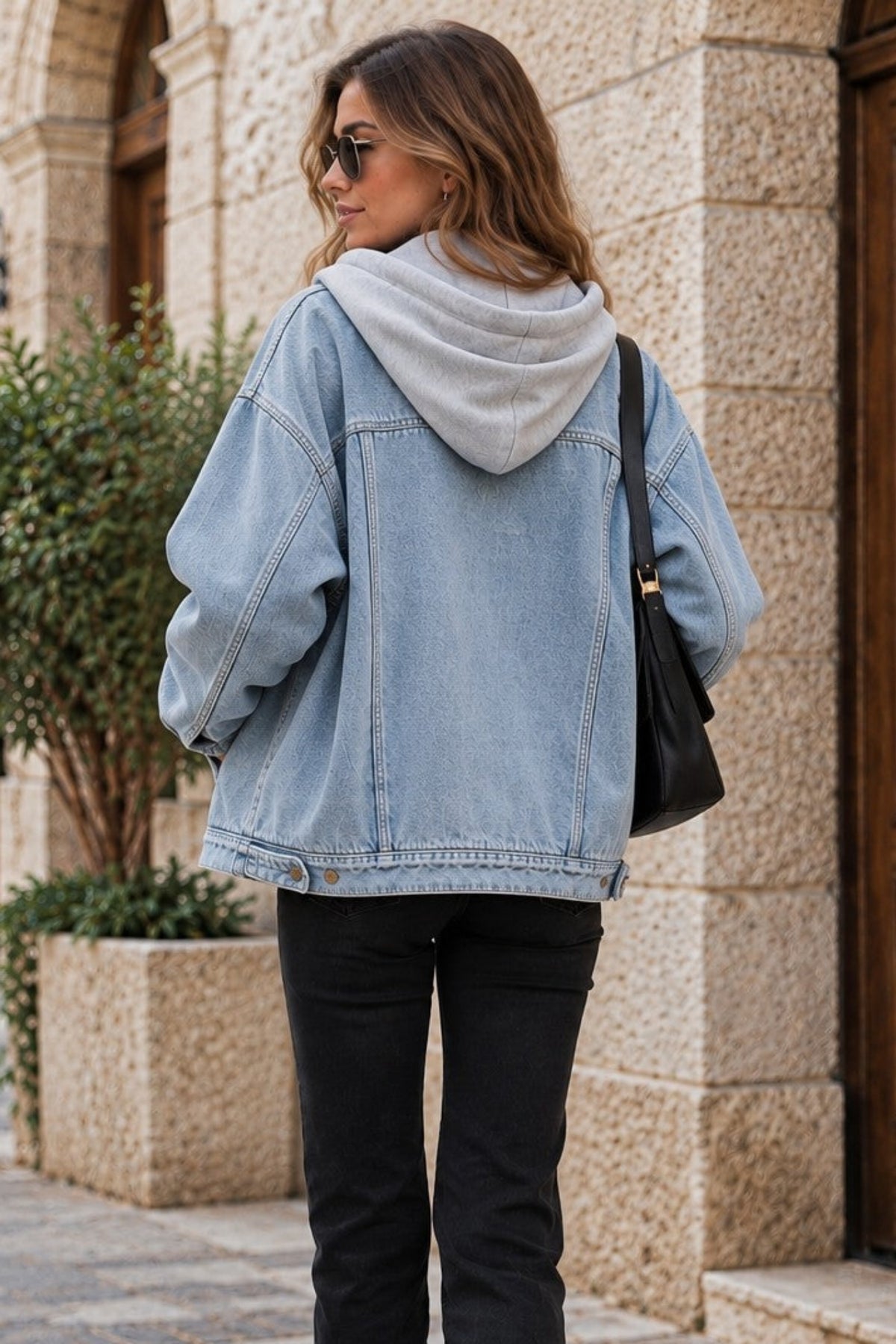 A woman walking on a cobblestone street wearing a light-wash blue denim jacket over a light gray hoodie, black flared pants, and carrying a black shoulder bag.