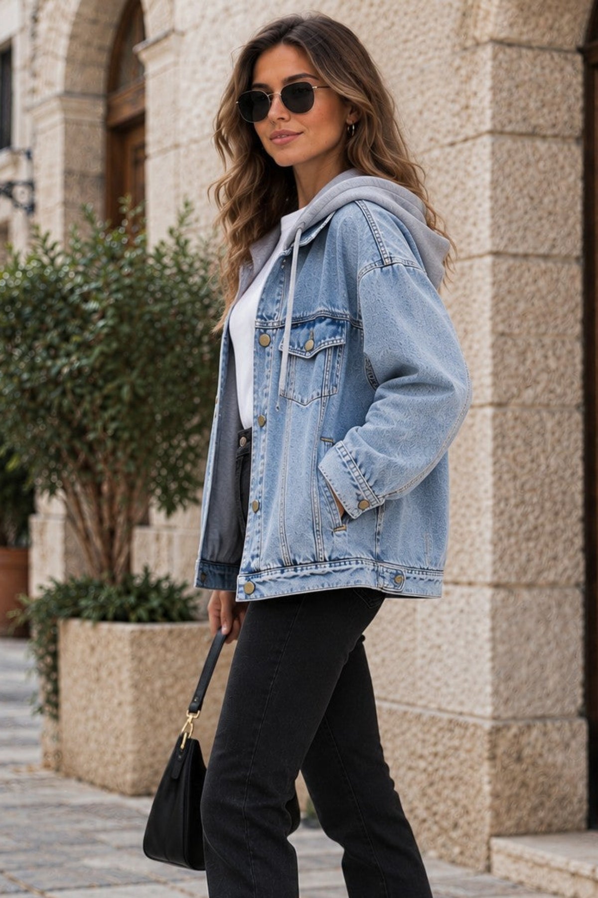 A woman stands on a cobblestone street wearing a light-wash blue denim jacket over a white top, black flared pants, white sneakers, and carrying a black handbag.
