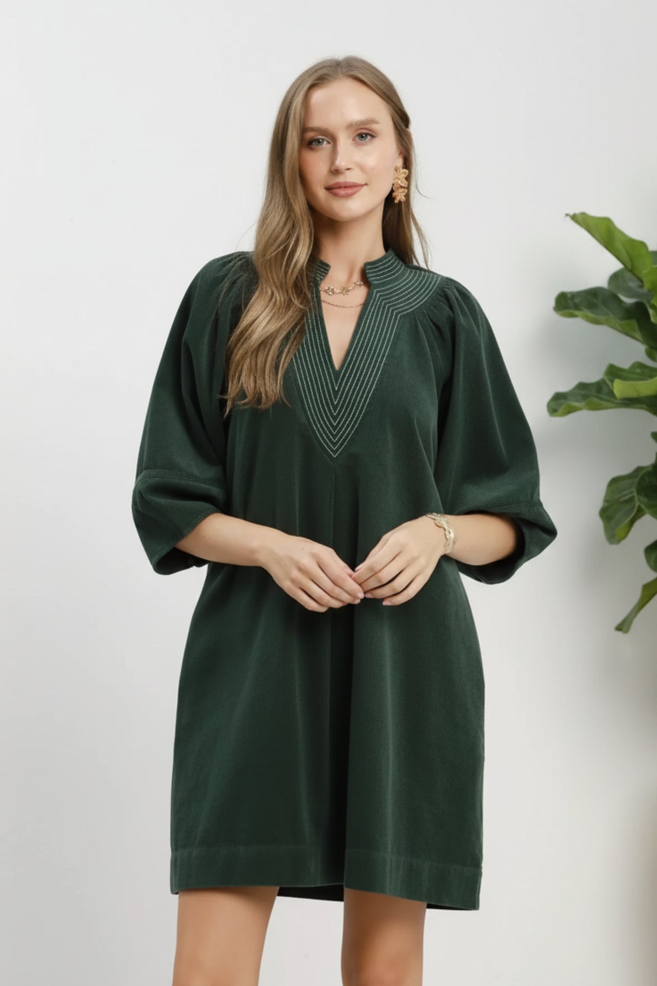 A woman modeling a dark green velvet-like tunic dress with a V-neckline and decorative embroidered detailing, shown against a white background