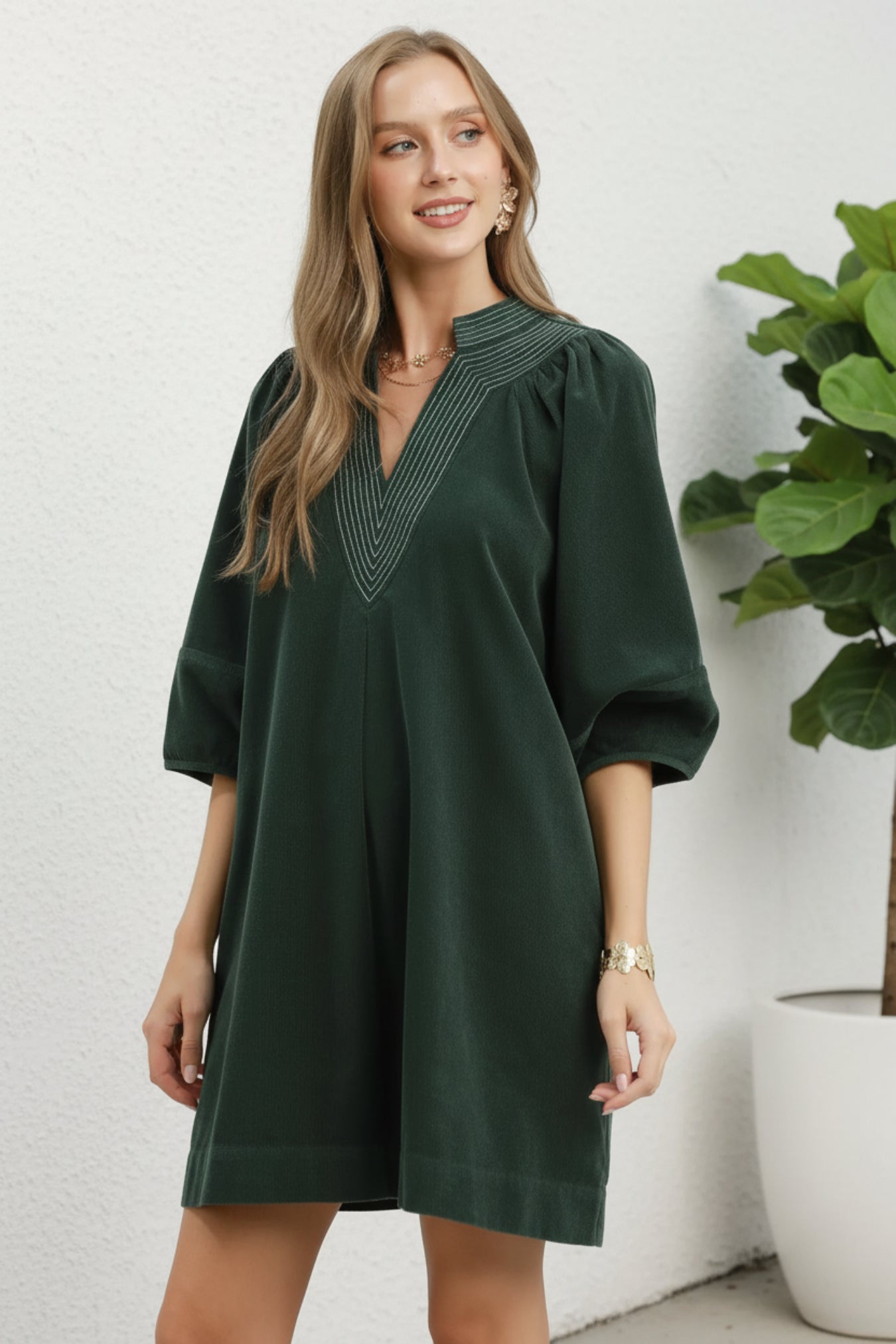 Woman wearing a hunter green dress with a white background next to a plant