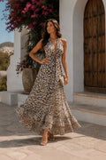 A woman models a sleeveless halter-neck maxi dress with an all-over leopard print pattern in beige, brown, and black. She is standing outdoors in front of a white stucco building with a rustic wooden door and potted plants.