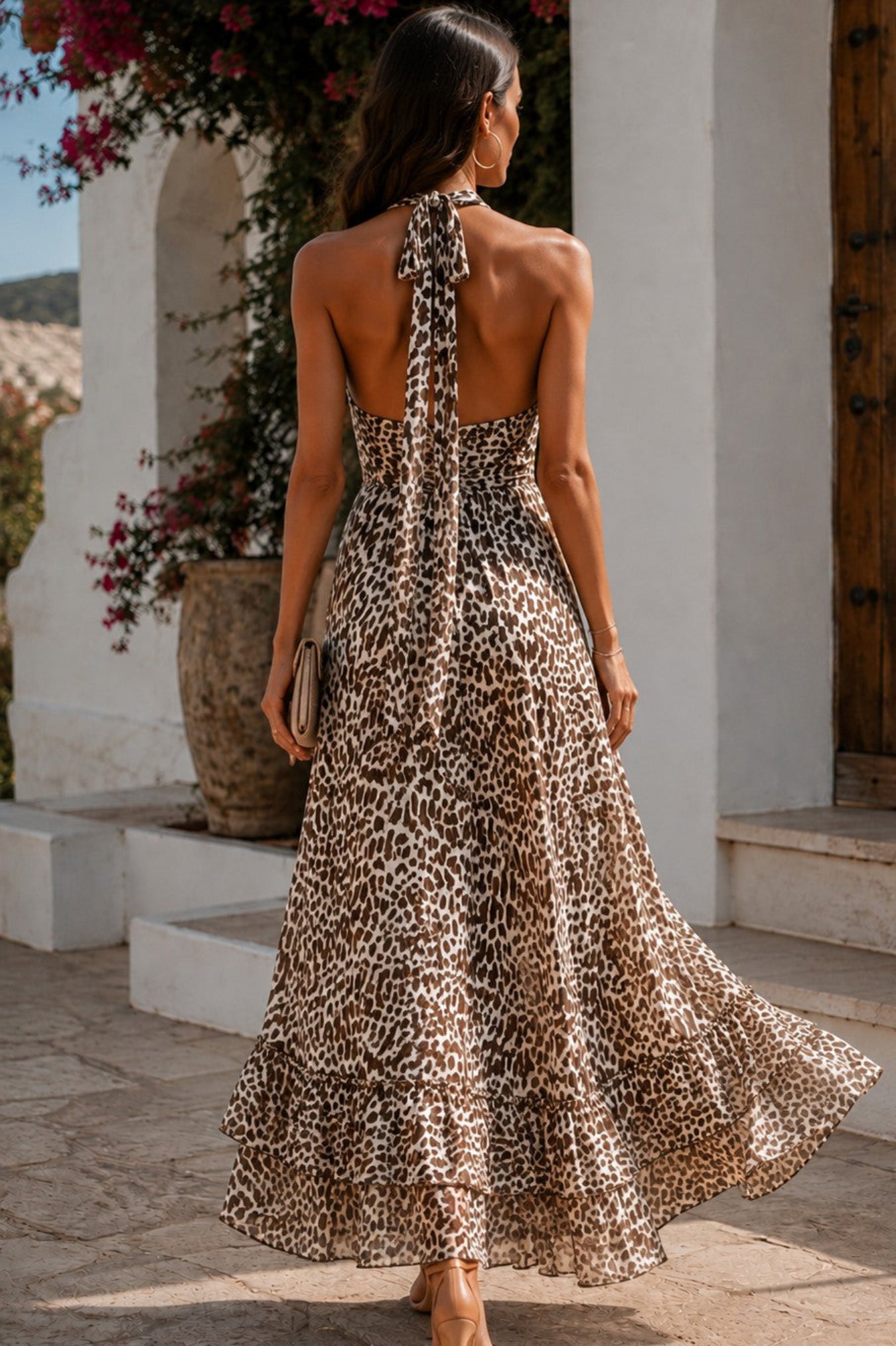 A woman wearing a leopard print halter maxi dress, photographed from behind as she walks outdoors in front of a white stucco building with a wooden door and pink flowers.