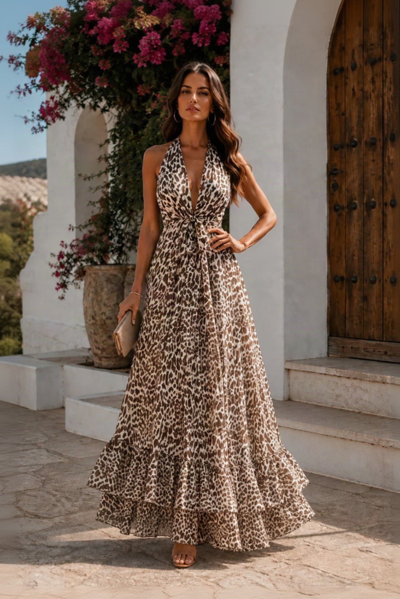 A woman models a sleeveless leopard-print maxi dress with a deep V-neckline, side tie detail, and a tiered ruffled hem, set against a Mediterranean-style stone patio and white stucco building background.
