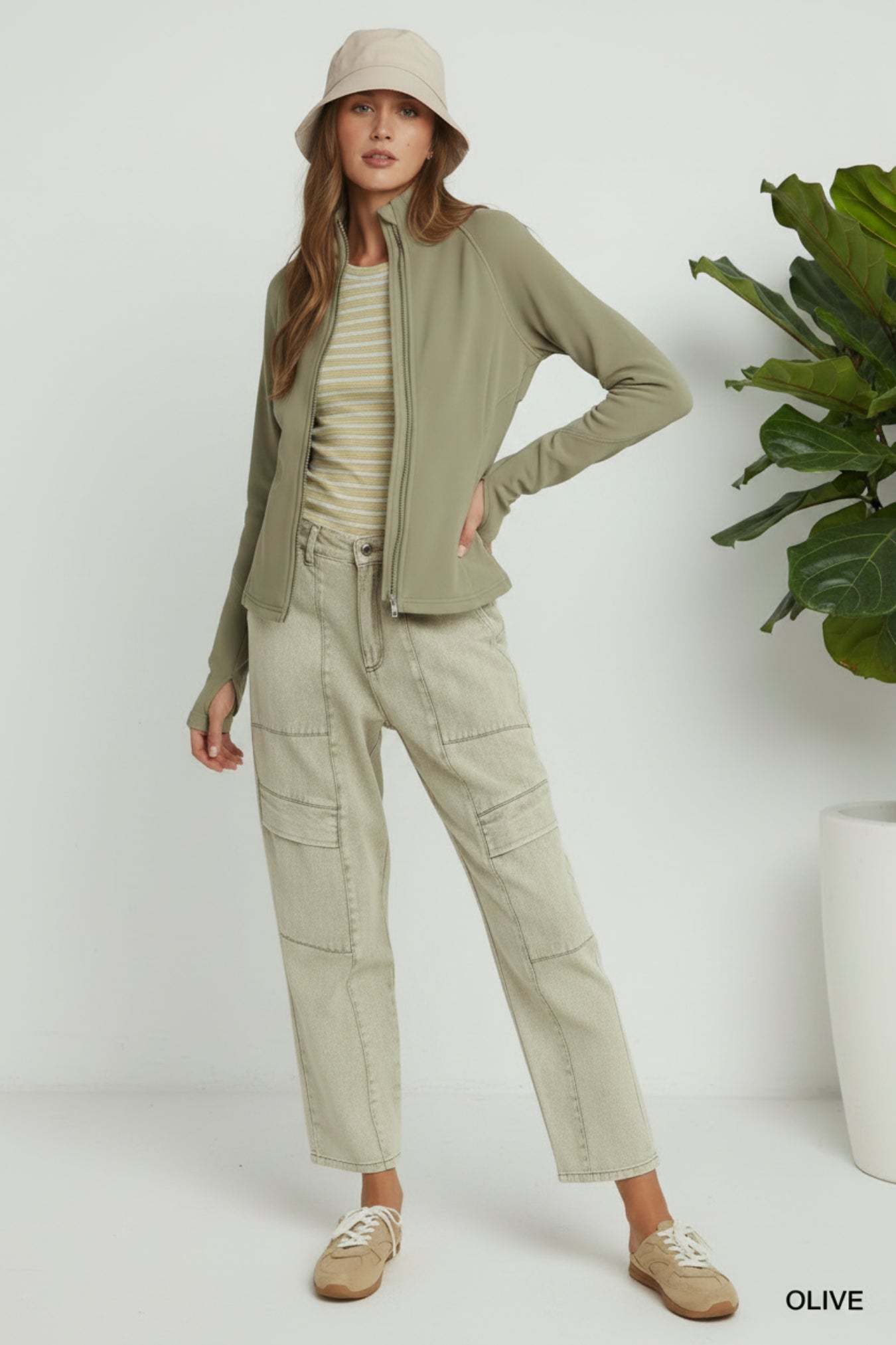 A model wearing an olive green zip-up cardigan, light beige cargo pants, a cream striped top, a beige bucket hat, and tan slip-on shoes, standing against a light gray studio background.