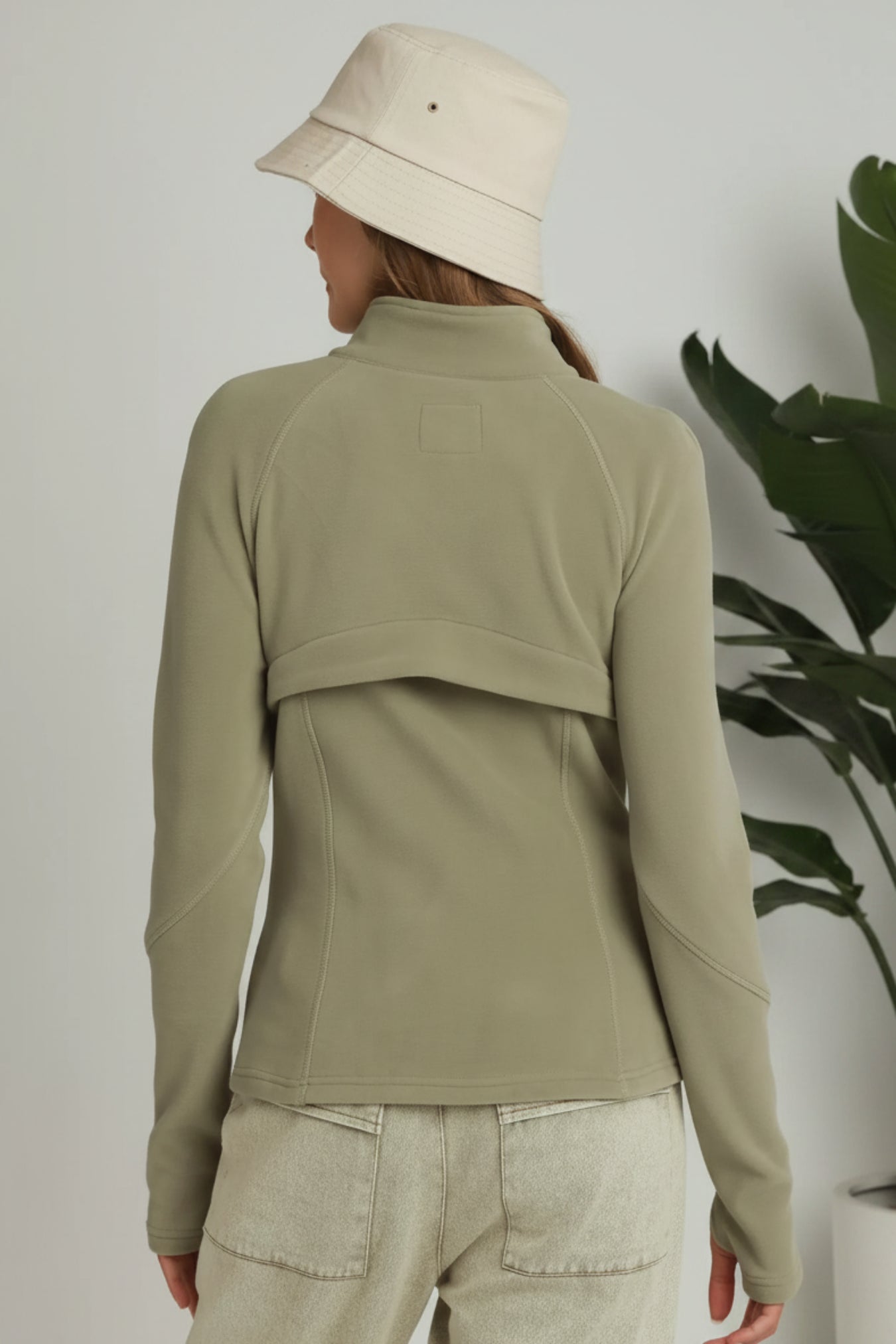 A woman shown from the back wearing an olive green long-sleeve athletic top with a high mock neck and a curved seam detail across the upper back, paired with light beige pants and a cream bucket hat against a white background.