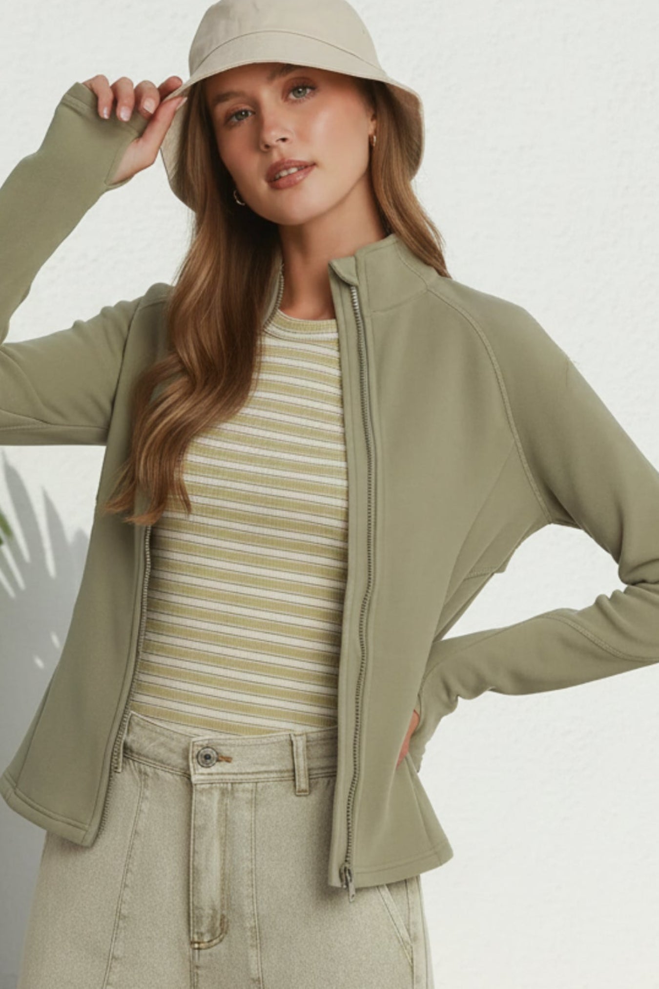 A woman modeling a light olive green zip-up jacket, paired with a beige and white striped t-shirt, beige cargo pants, and a matching beige bucket hat, standing against a white wall.
