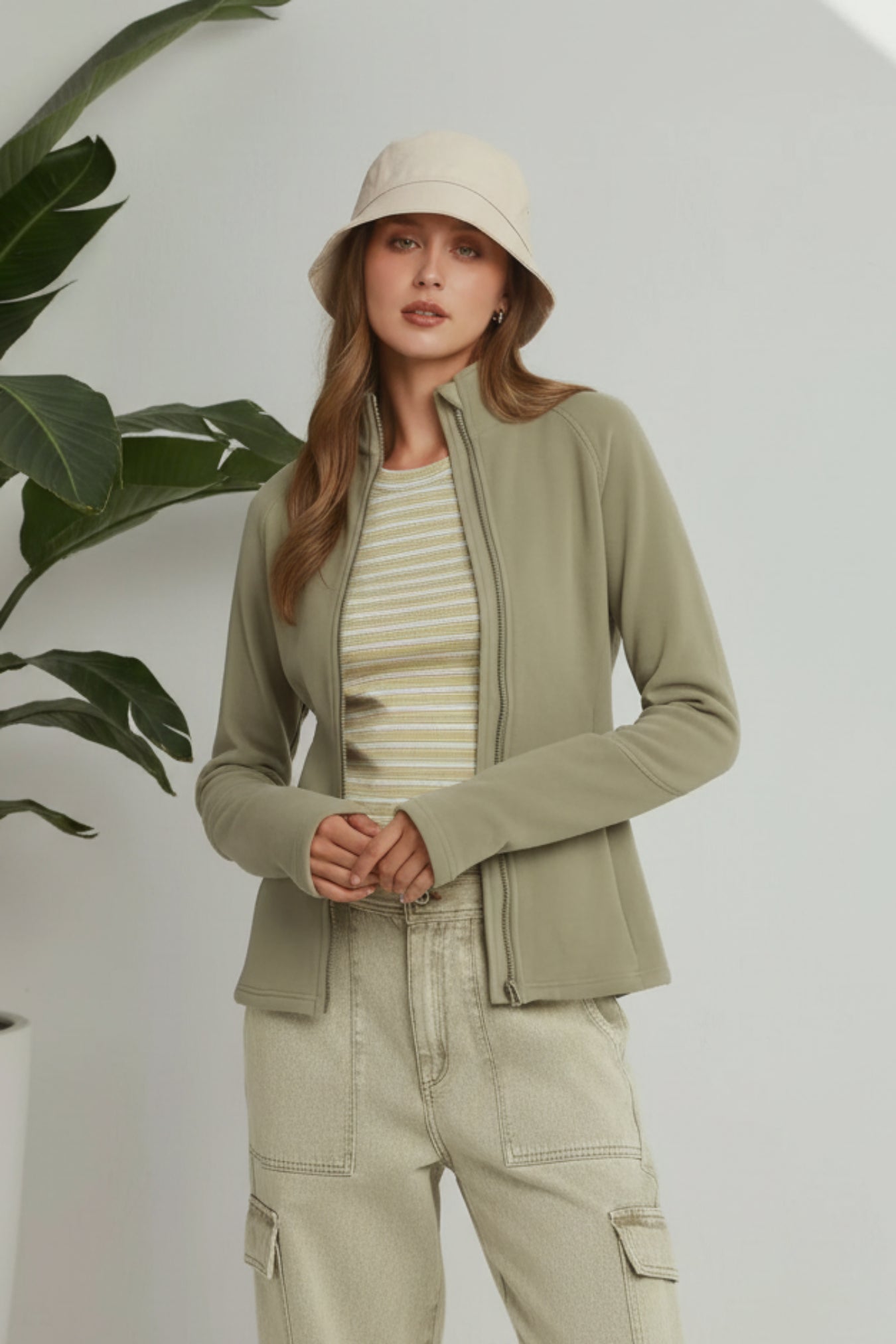 A woman models a light olive green zip-up jacket over a cream and beige striped t-shirt, paired with beige cargo pants and a matching beige bucket hat, set against a light gray wall with a green plant leaf on the left.