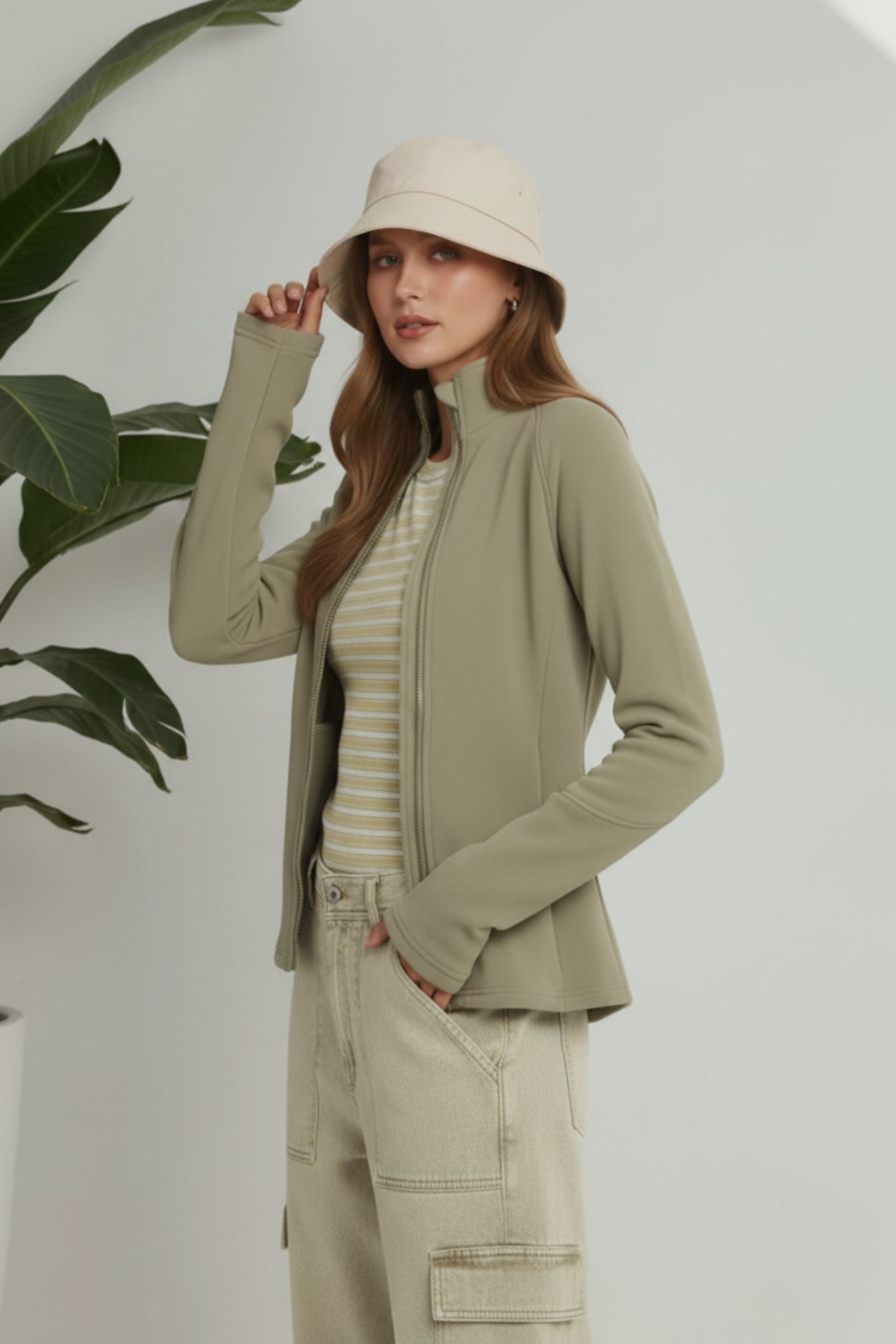 A woman models a light olive green zip-up jacket over a cream and beige striped t-shirt, paired with beige cargo pants and a matching beige bucket hat, set against a white wall with a green plant leaf on the left.