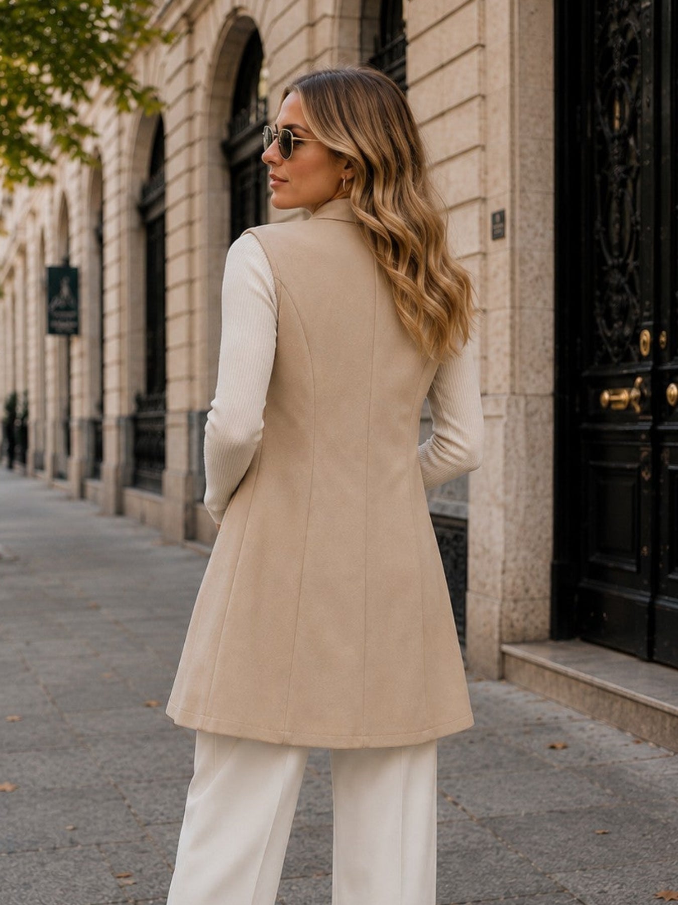 A woman stands on a city sidewalk wearing a beige sleeveless tailored vest over a white long-sleeve top and white wide-leg pants, with tan heels and sunglasses.