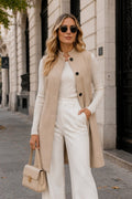A woman stands on a city sidewalk wearing a beige sleeveless coat over a cream top and white wide-leg pants, holding a beige handbag.