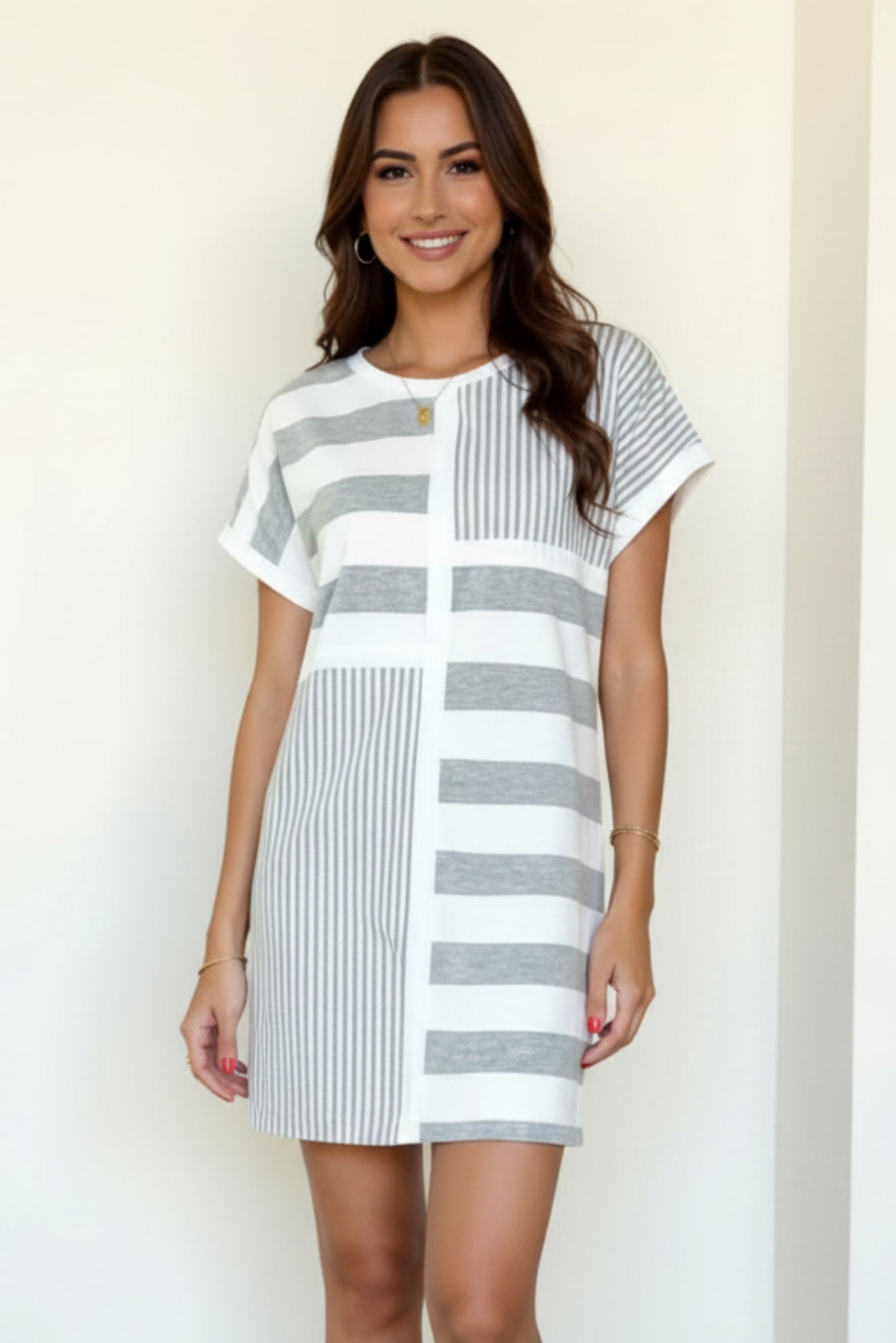 A woman modeling a short-sleeve mini dress with a gray and white striped patchwork pattern, paired with white slip-on sneakers, standing against a minimalist off-white wall.