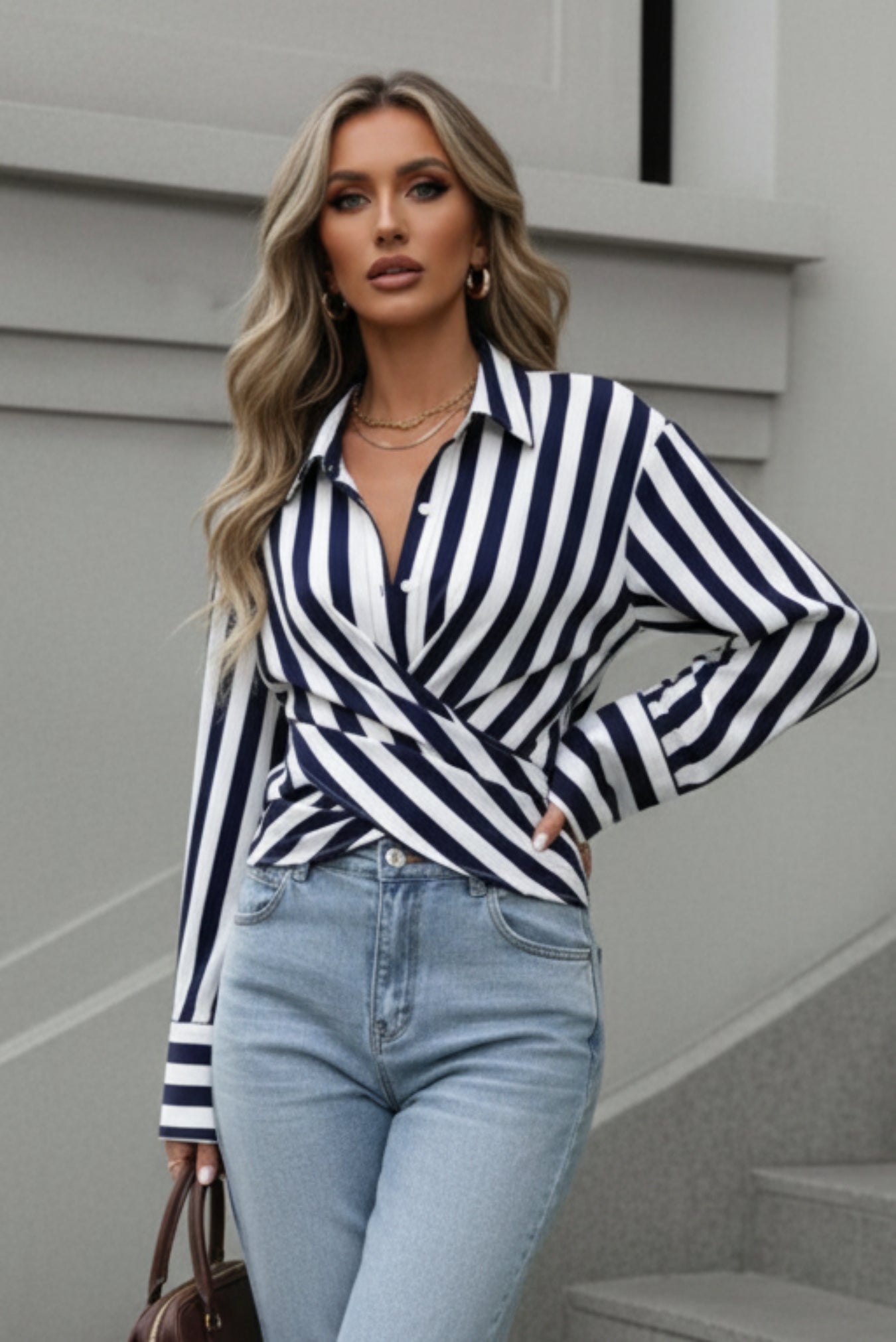 A woman walking outdoors in front of gray stone steps, wearing a navy and white vertically striped wrap-style long-sleeve blouse, light blue jeans, white sneakers, and carrying a brown handbag.