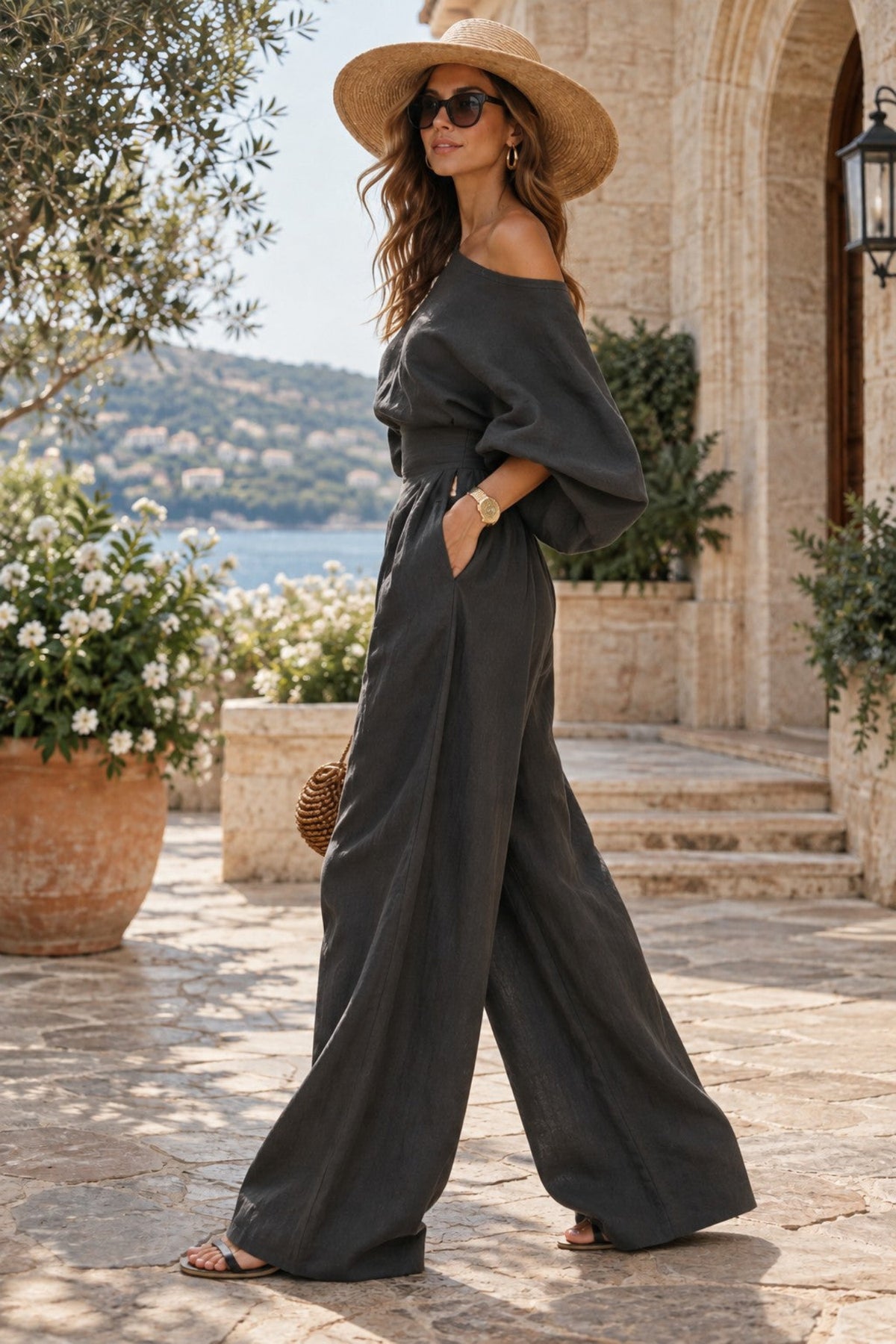 A woman wearing a dark charcoal gray off-the-shoulder wide-leg jumpsuit, a straw sun hat, and sunglasses, posing outdoors on a stone patio with a scenic background of a villa and sea.