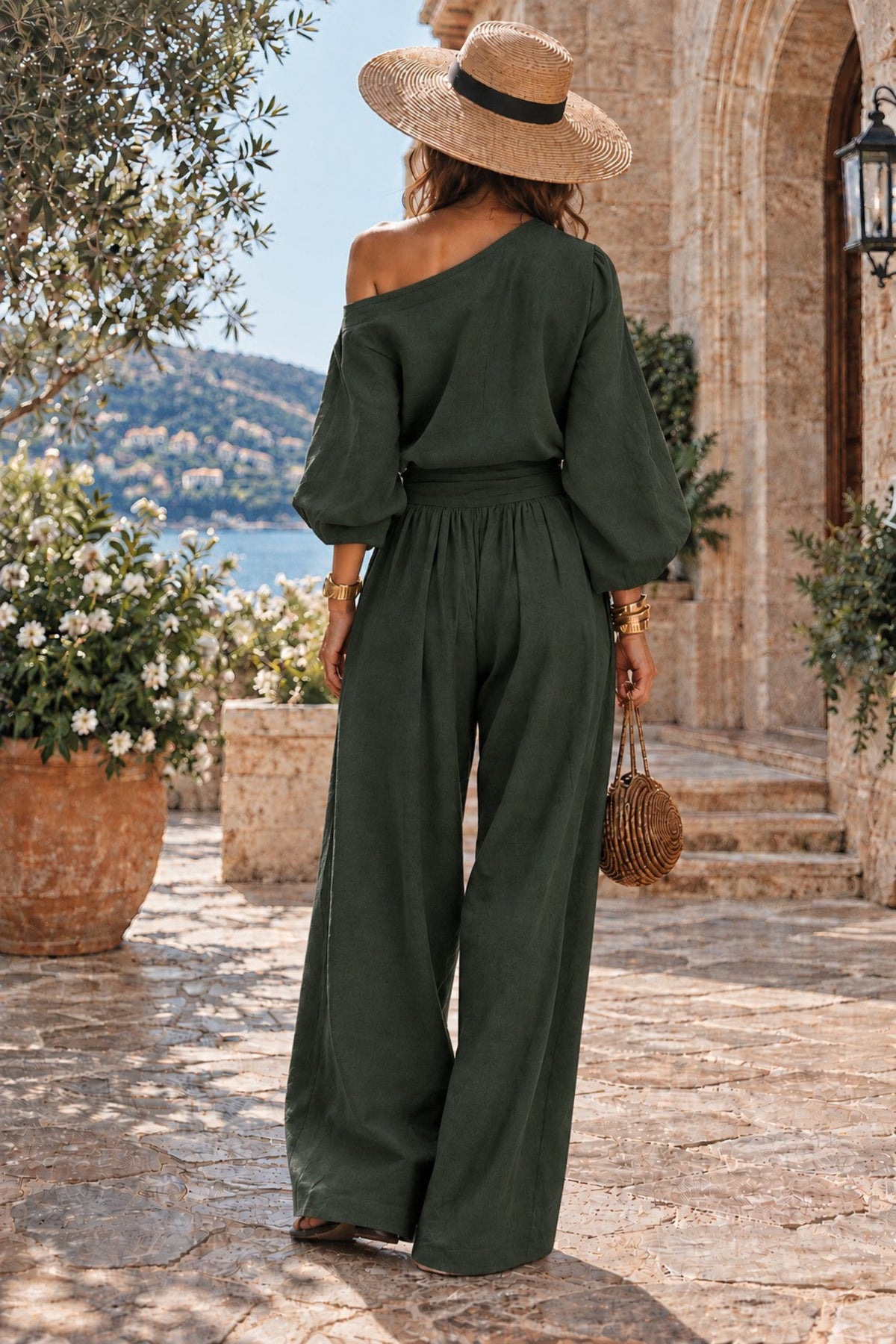 A woman stands on a stone walkway outdoors, shown from the back, wearing an olive green off-the-shoulder wide-leg jumpsuit, a wide-brim straw hat, and holding a small woven bag.