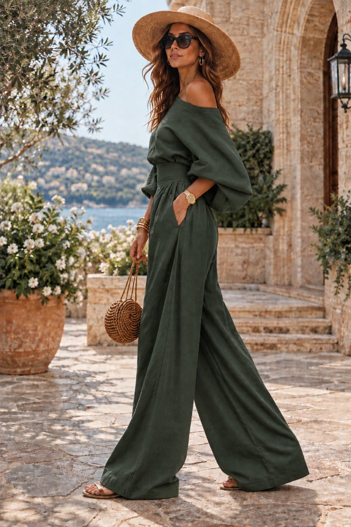 A woman wearing an olive green off-the-shoulder wide-leg jumpsuit, a straw hat, and sunglasses walks outdoors on a stone patio near a building with a sea view in the background.