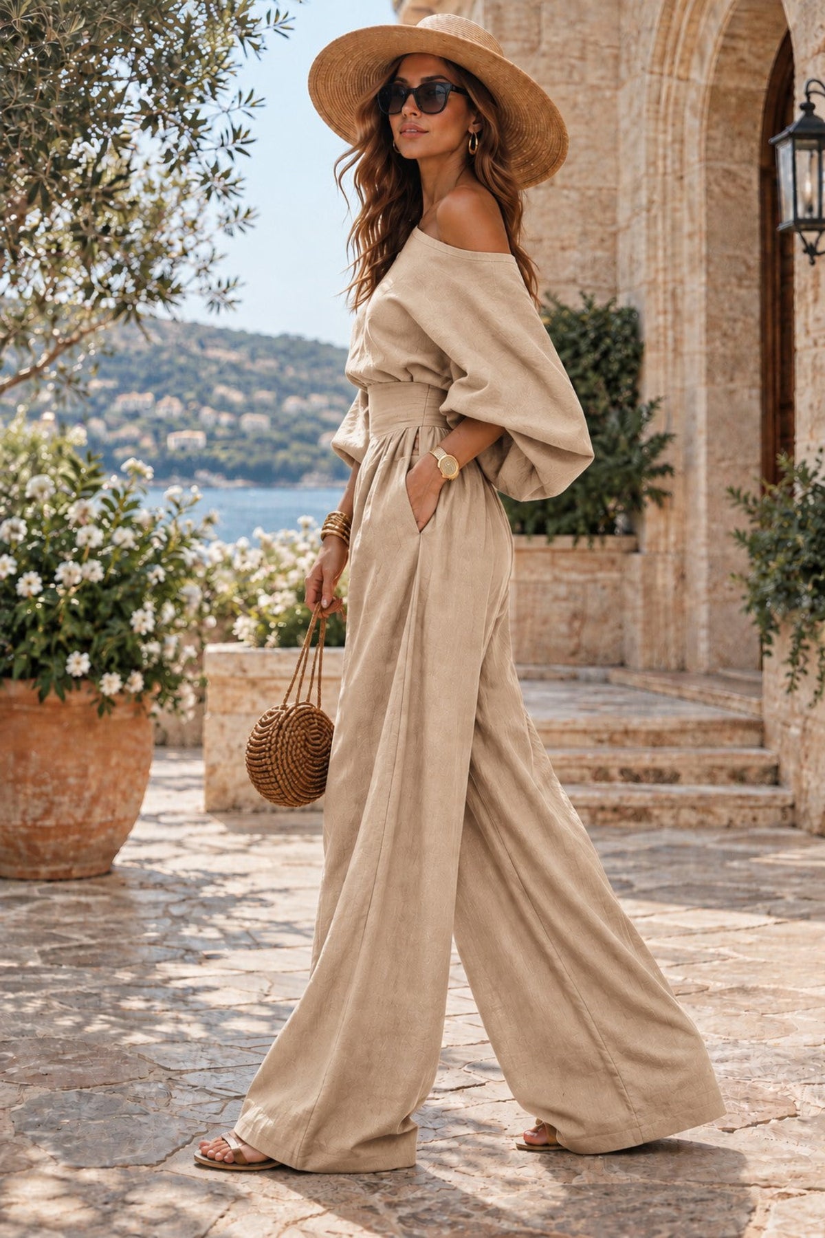 A woman wearing a beige off-the-shoulder wide-leg jumpsuit, a straw hat, and sunglasses walks outdoors in a sunlit stone courtyard with potted plants and a distant body of water.