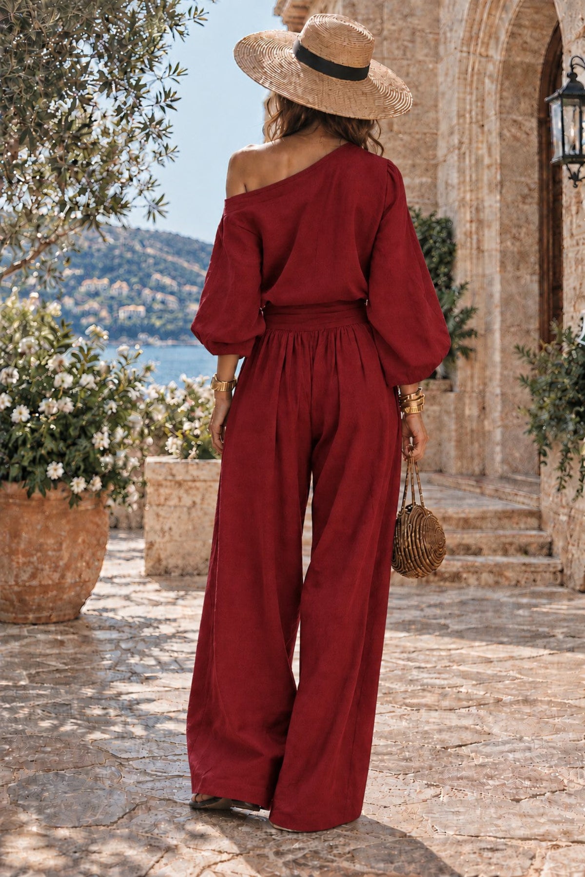 A woman stands outdoors on a stone walkway wearing a deep burgundy one-shoulder jumpsuit with balloon sleeves and wide legs, paired with a wide-brim straw hat and carrying a small woven bag.