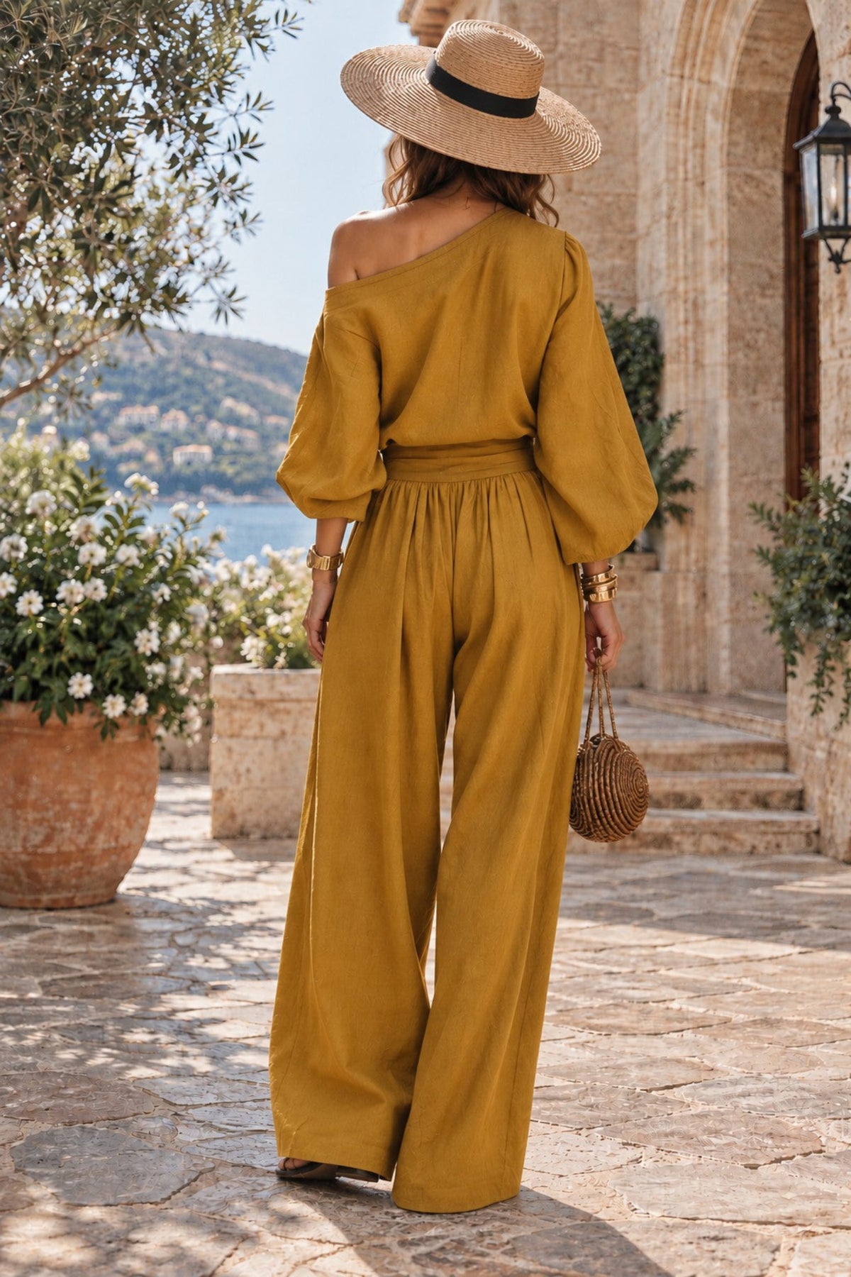 A woman stands on a stone walkway outdoors wearing a mustard yellow one-shoulder jumpsuit with wide legs, a straw hat, and carrying a small woven bag.