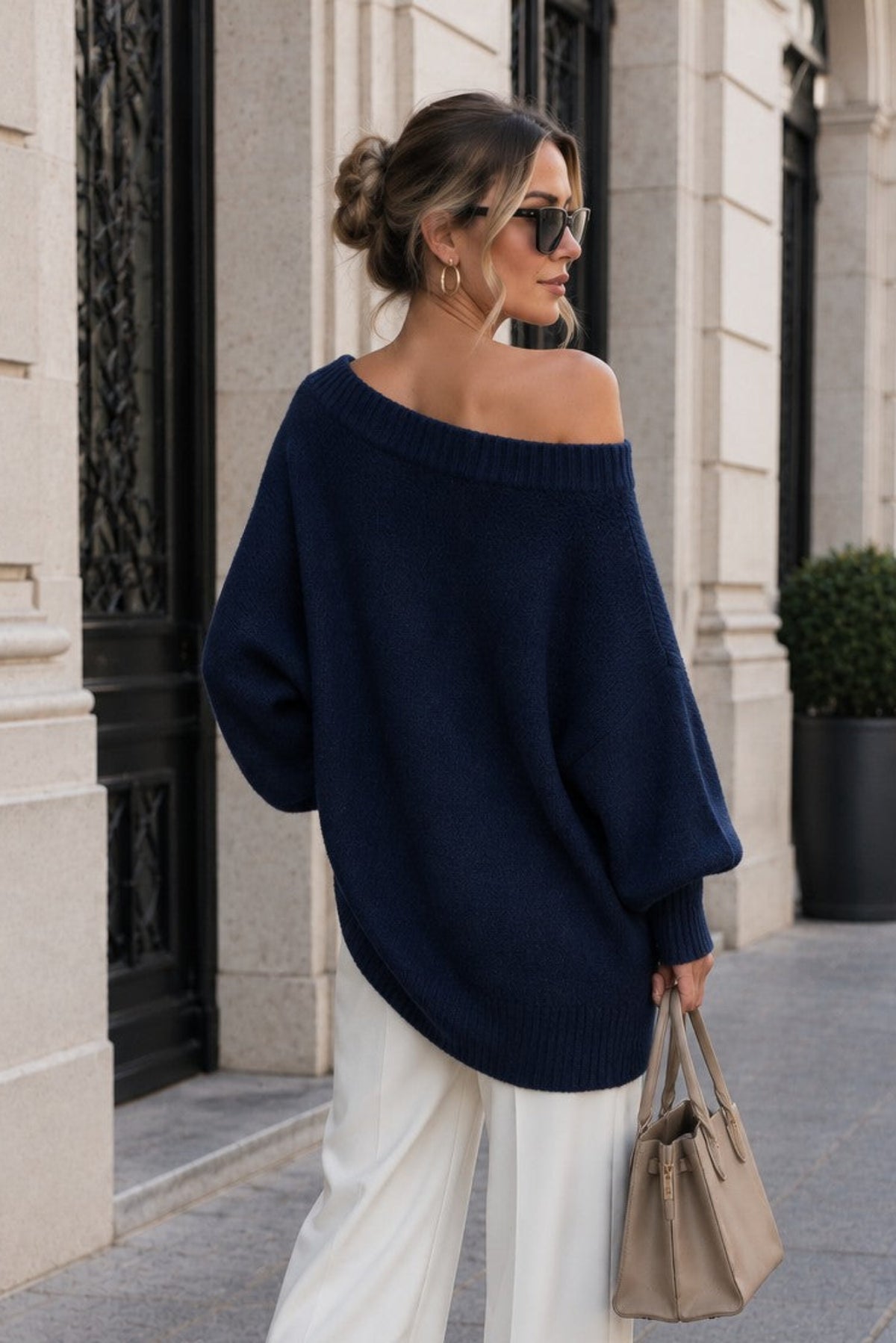 A woman stands on a city sidewalk wearing an oversized off-the-shoulder navy sweater, white wide-leg pants, and carrying a beige handbag.