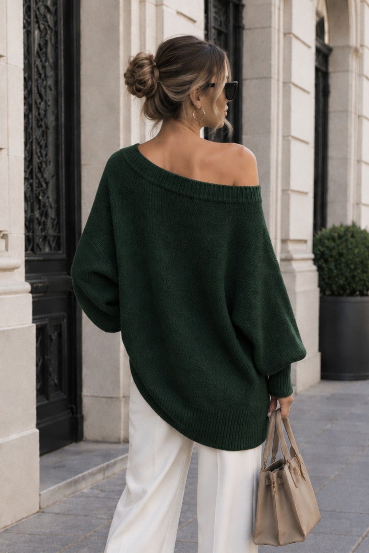 A woman stands on a city sidewalk wearing an oversized dark green off-the-shoulder knit sweater, white wide-leg pants, and carrying a beige handbag.