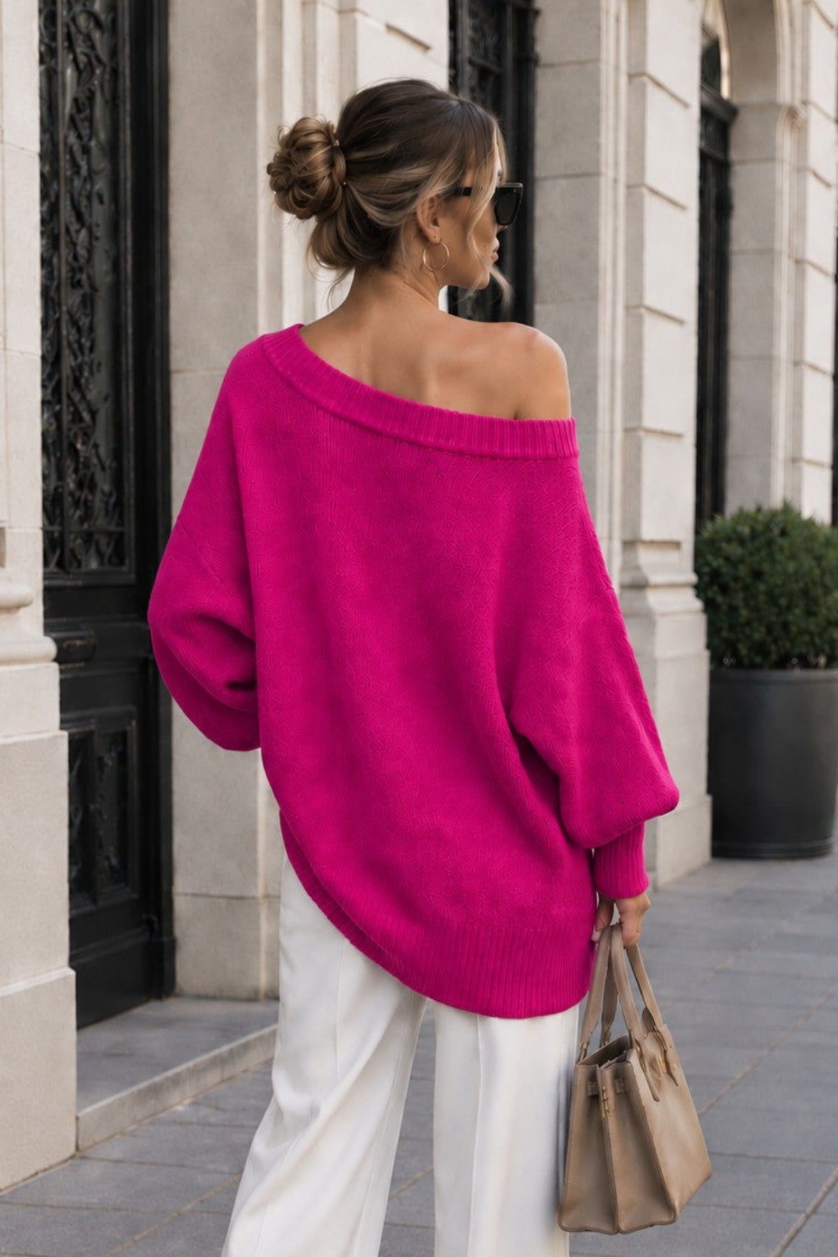 A woman stands on a city sidewalk wearing an oversized off-the-shoulder bright pink sweater, white wide-leg pants, and carrying a tan handbag.