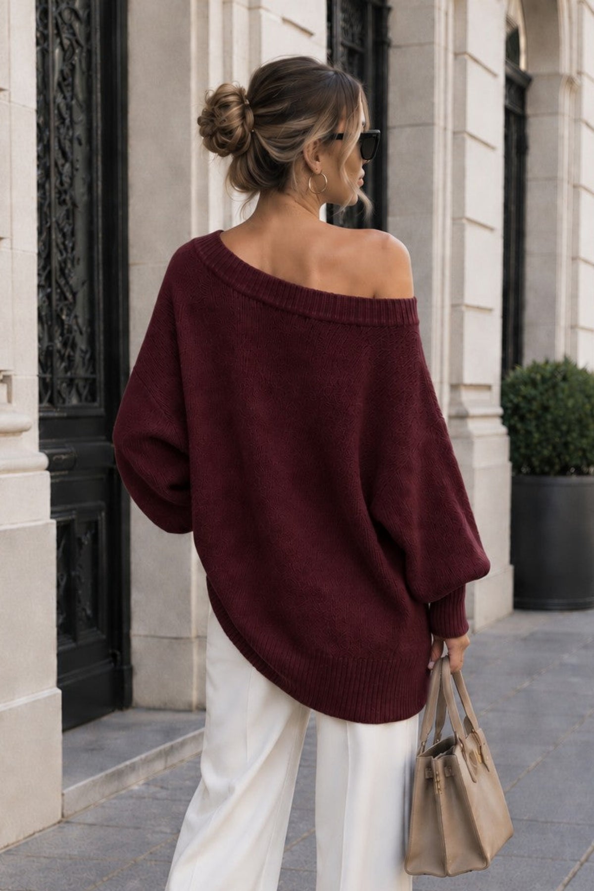 A woman photographed from behind wearing a loose off-the-shoulder burgundy sweater and white wide-leg pants, standing on a stone sidewalk in front of a building with columns and potted plants.