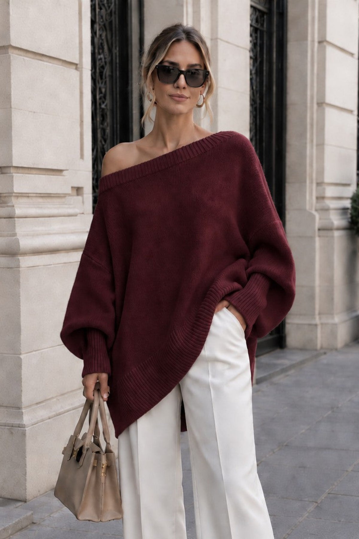A woman stands on a city sidewalk wearing an oversized off-the-shoulder burgundy knit sweater, white wide-leg pants, and beige pointed shoes, holding a taupe handbag.