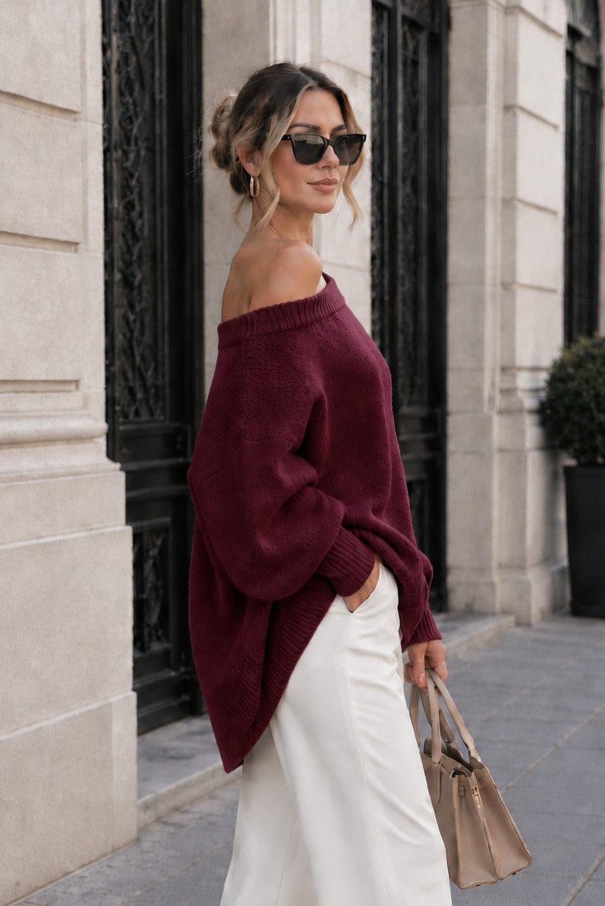A woman stands on a city sidewalk wearing an off-the-shoulder burgundy knit sweater and white wide-leg trousers, holding a beige handbag. The background shows a stone building facade with dark metal doors.