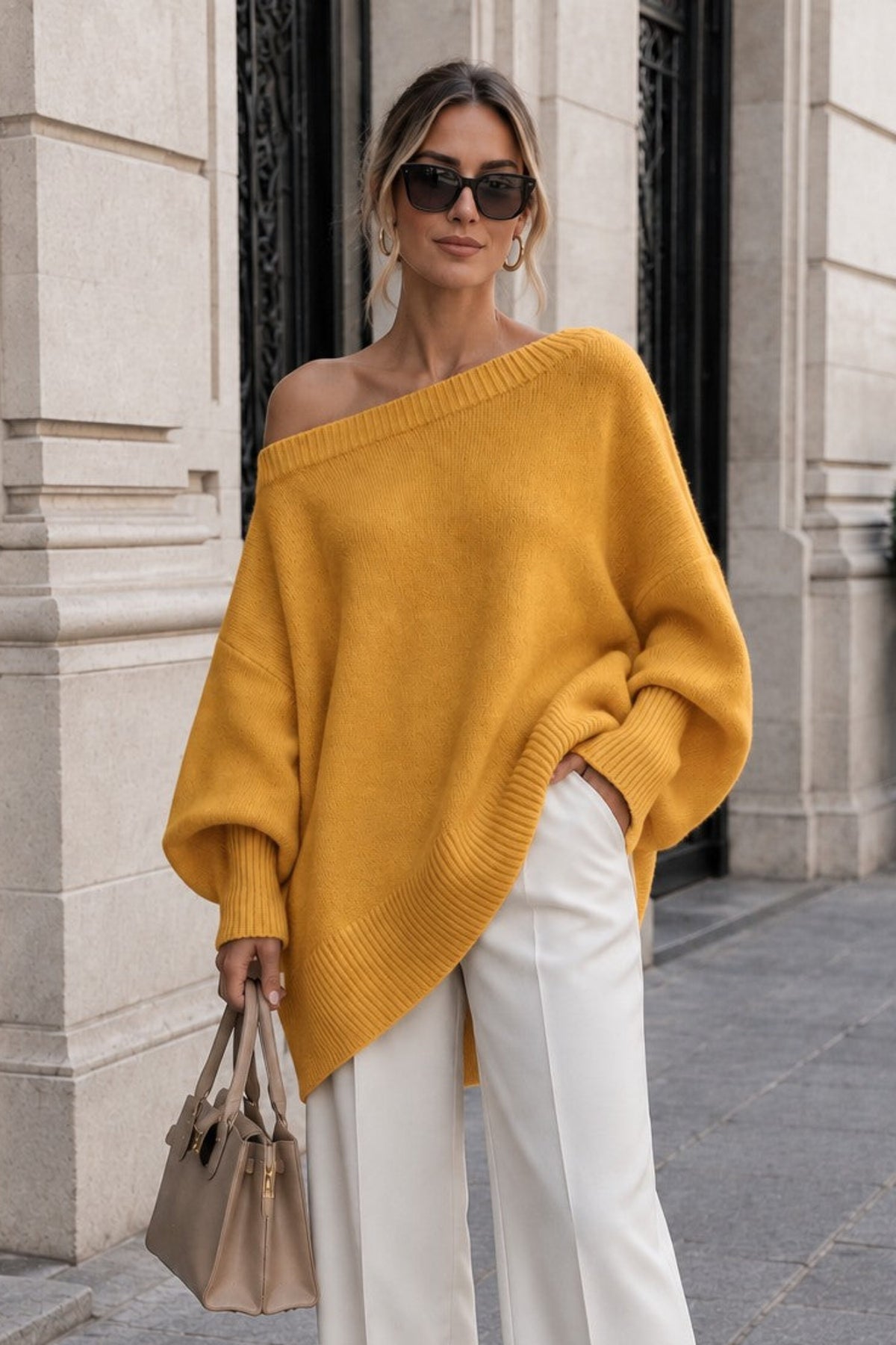 A woman stands on a city sidewalk wearing a mustard yellow off-the-shoulder knit sweater, white wide-leg pants, beige pointed shoes, and holding a beige handbag.