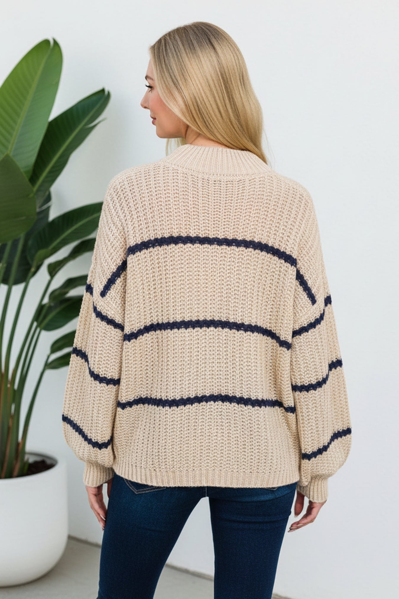 A woman wearing a chunky knit sweater with horizontal navy blue stripes on a warm beige/taupe base, paired with dark blue jeans, shown from a back-three-quarter angle against a white background with a plant on the side.