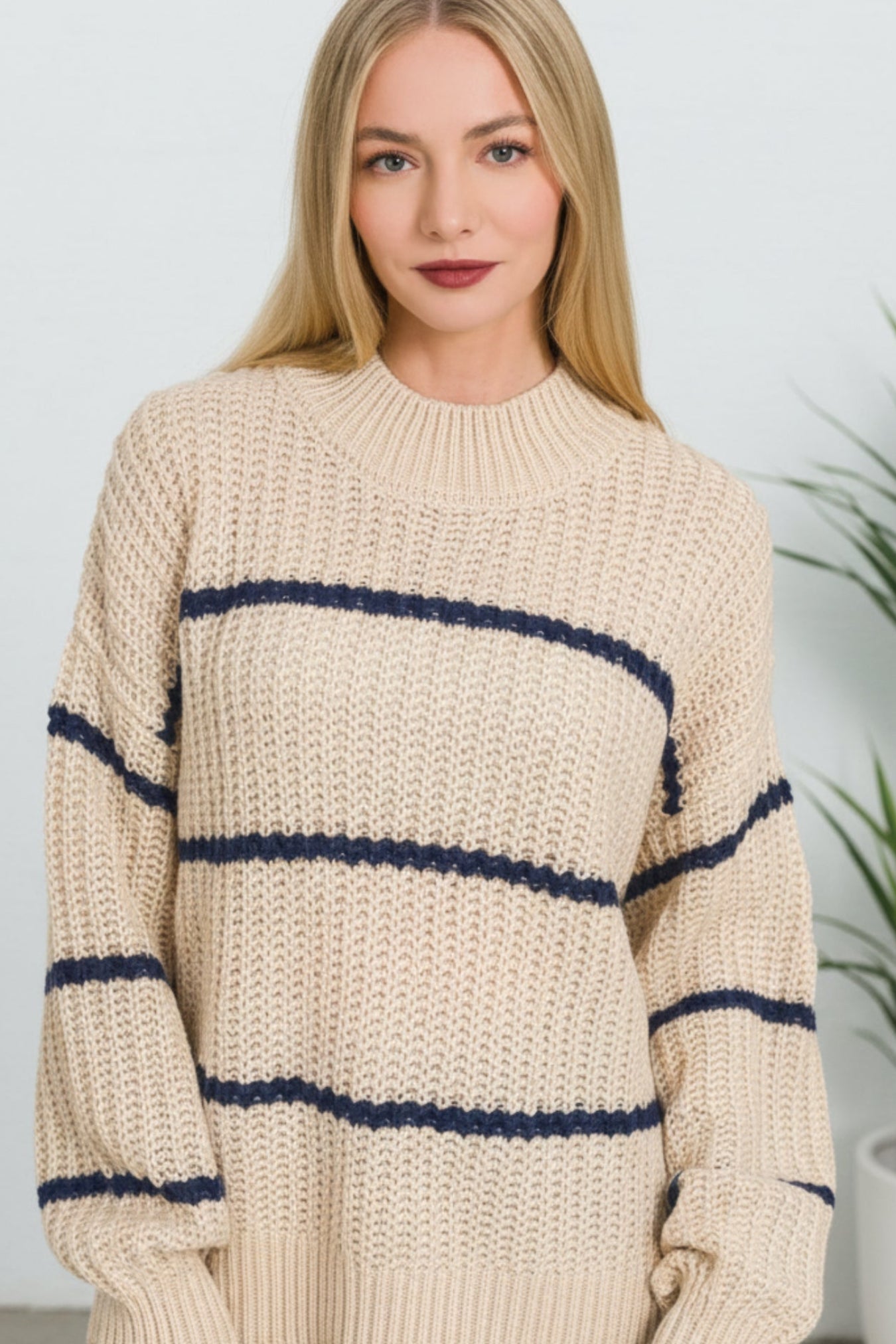 A woman wearing a chunky knit sweater in cream with horizontal navy stripes, photographed against a light gray indoor background with a blurred plant visible on the right.
