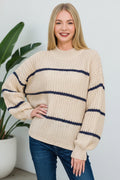 A woman wearing a chunky knit crewneck sweater in taupe with navy blue horizontal stripes, paired with blue jeans, against a plain white background.