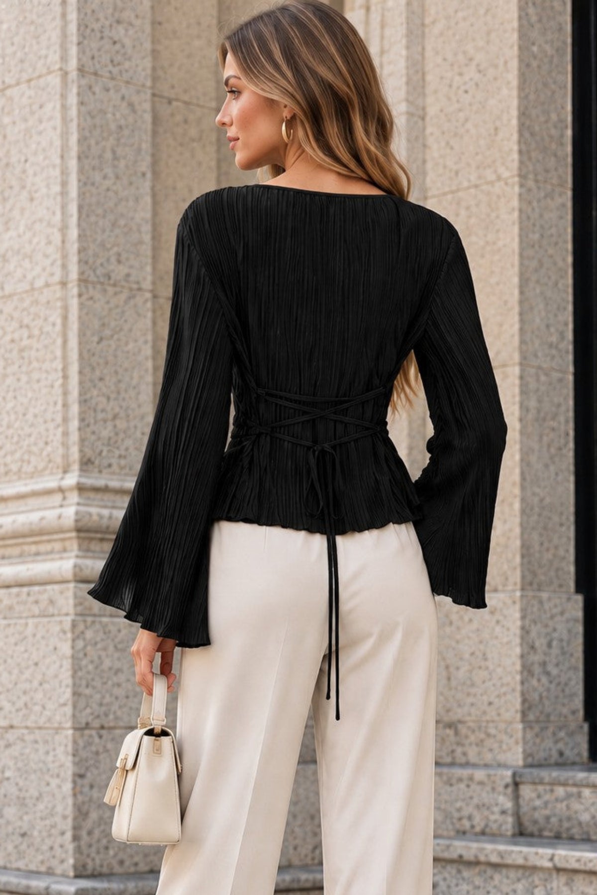 A woman photographed from the back wearing a black pleated long-sleeve top with a tied waist and cream wide-leg pants, standing outdoors in front of a stone building.